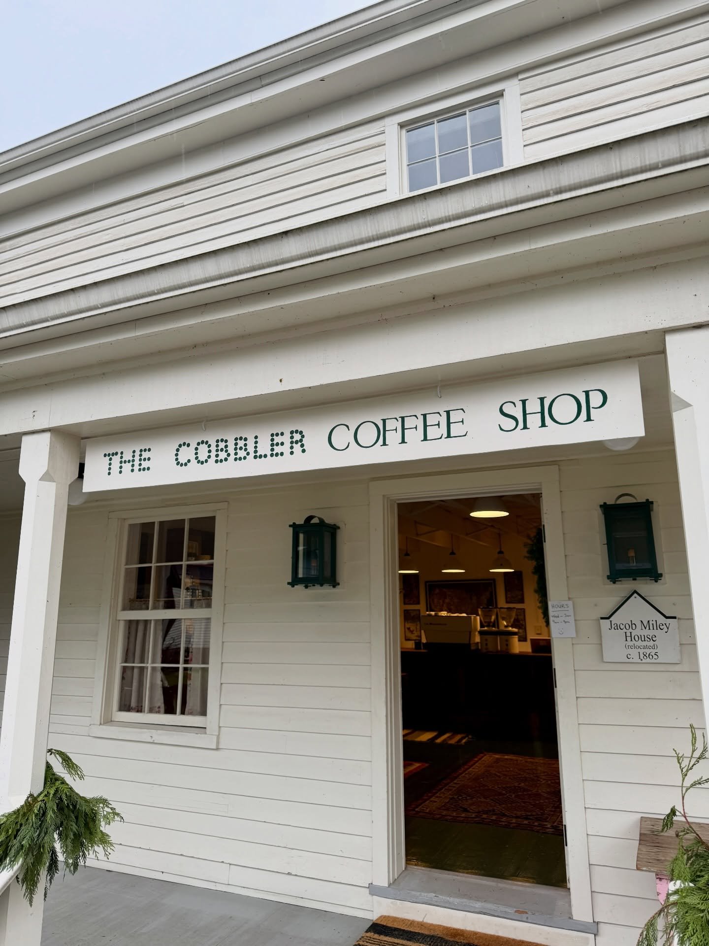 The Cobbler Coffee Shop, a white wooden building with an open door, hanging lanterns, and a sign showing it is the Jacob Miley House, relocated around 1865.