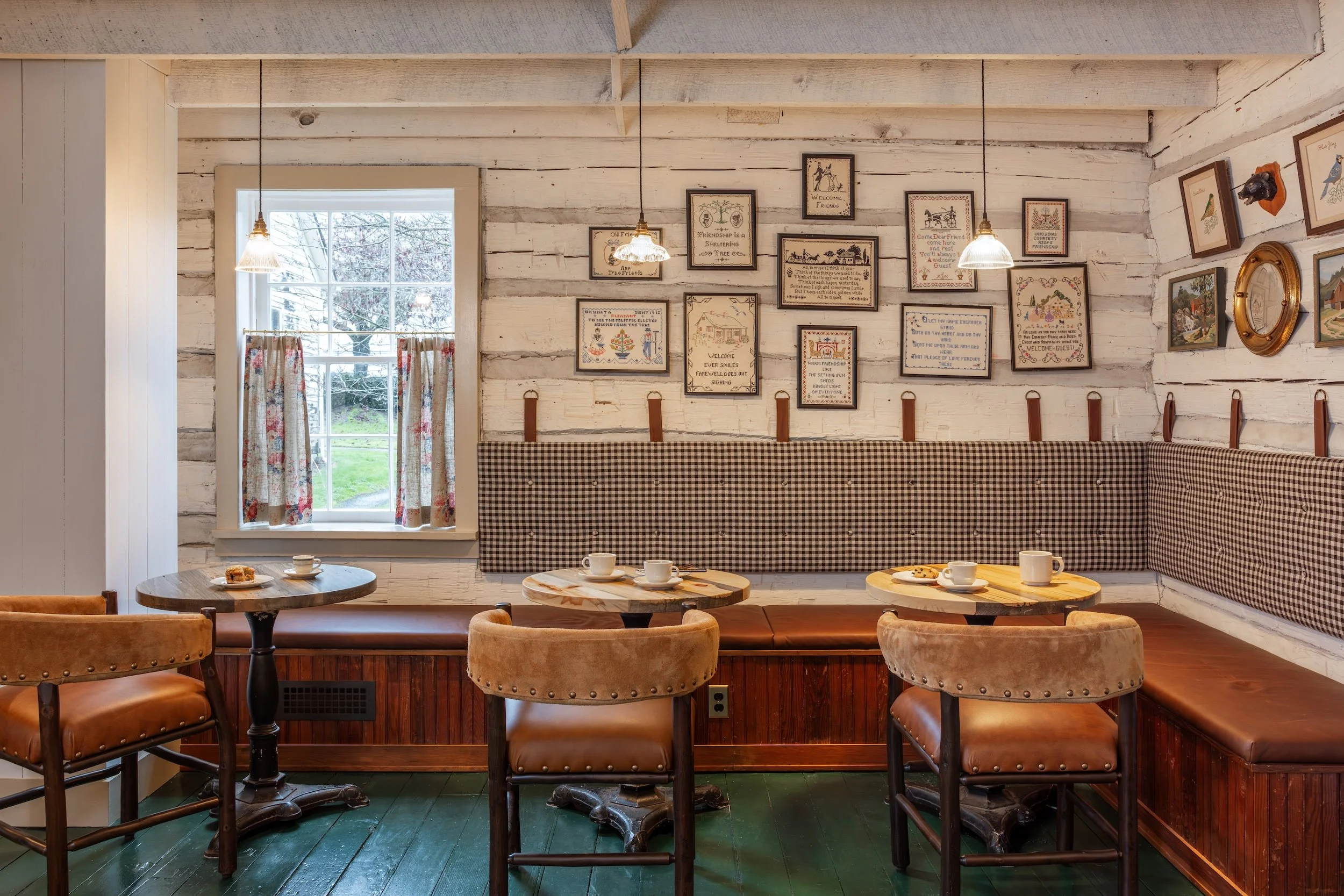 Cozy cafe interior with wooden tables, cushioned chairs, framed wall art, window with floral curtains, and a corner bench with checkered upholstery.
