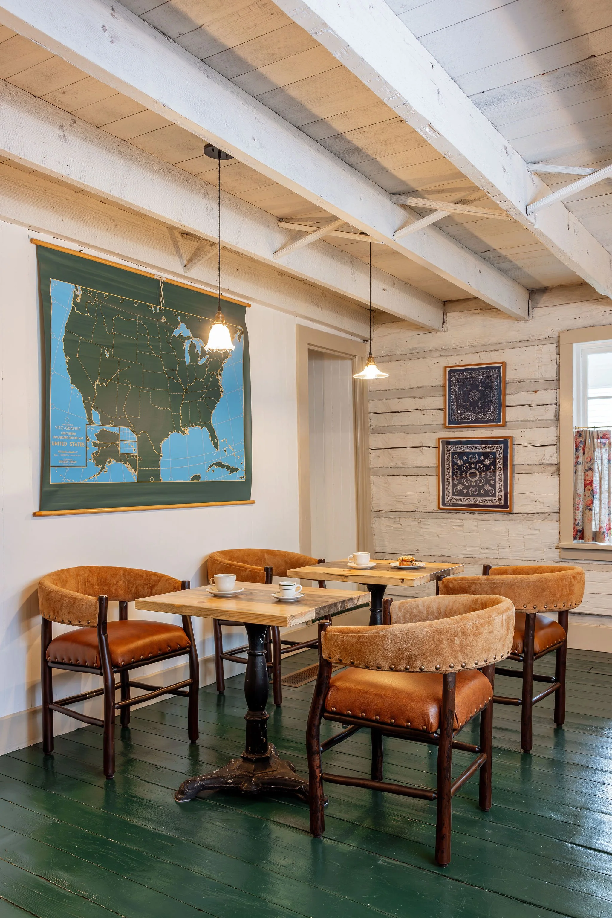 A cozy dining area with four brown upholstered chairs around two wooden tables, set with mugs and plates, in a rustic room with whitewashed wooden walls, green wooden flooring, a map of the United States on the wall, two hanging pendant lights, and f