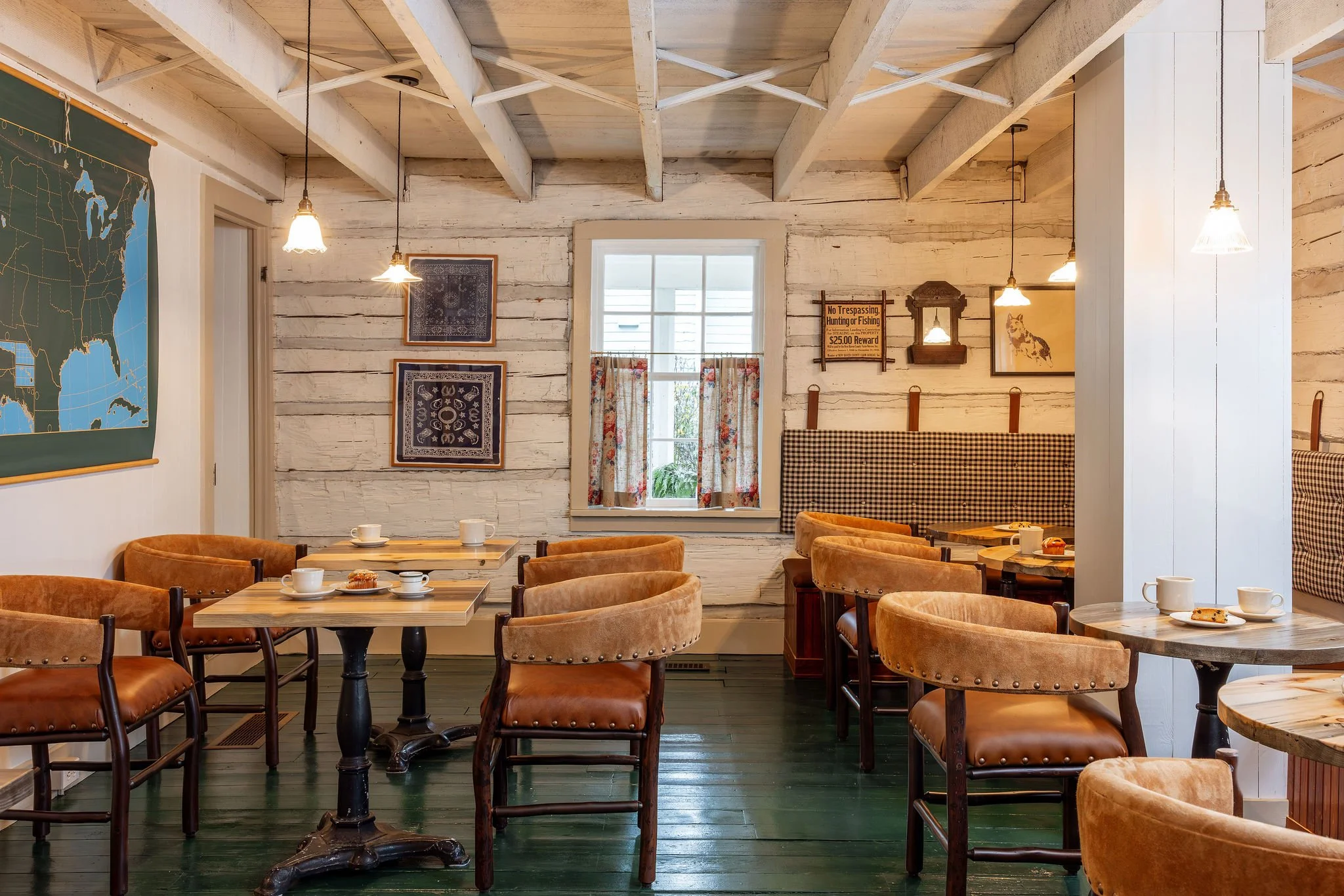 Interior of a cozy café with whitewashed wooden walls, framed artwork, and vintage-style lighting. There are several round and rectangular wooden tables with tan upholstered chairs. Cups and plates are on the tables, and a window with floral curtains