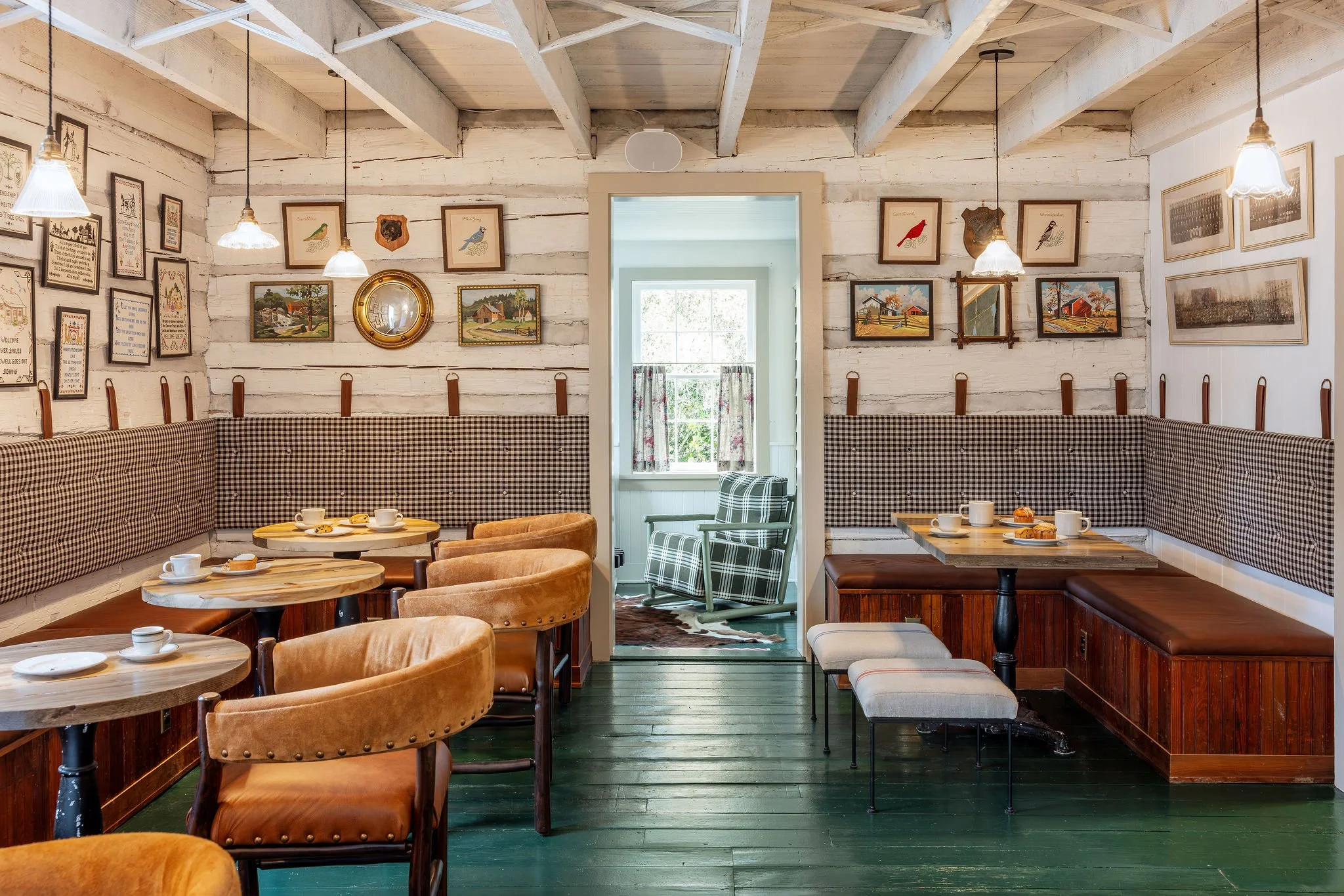Cozy cafe interior with white wooden walls, framed artwork, checkered and plaid accents, wooden tables, and chairs with cups and plates set for breakfast.