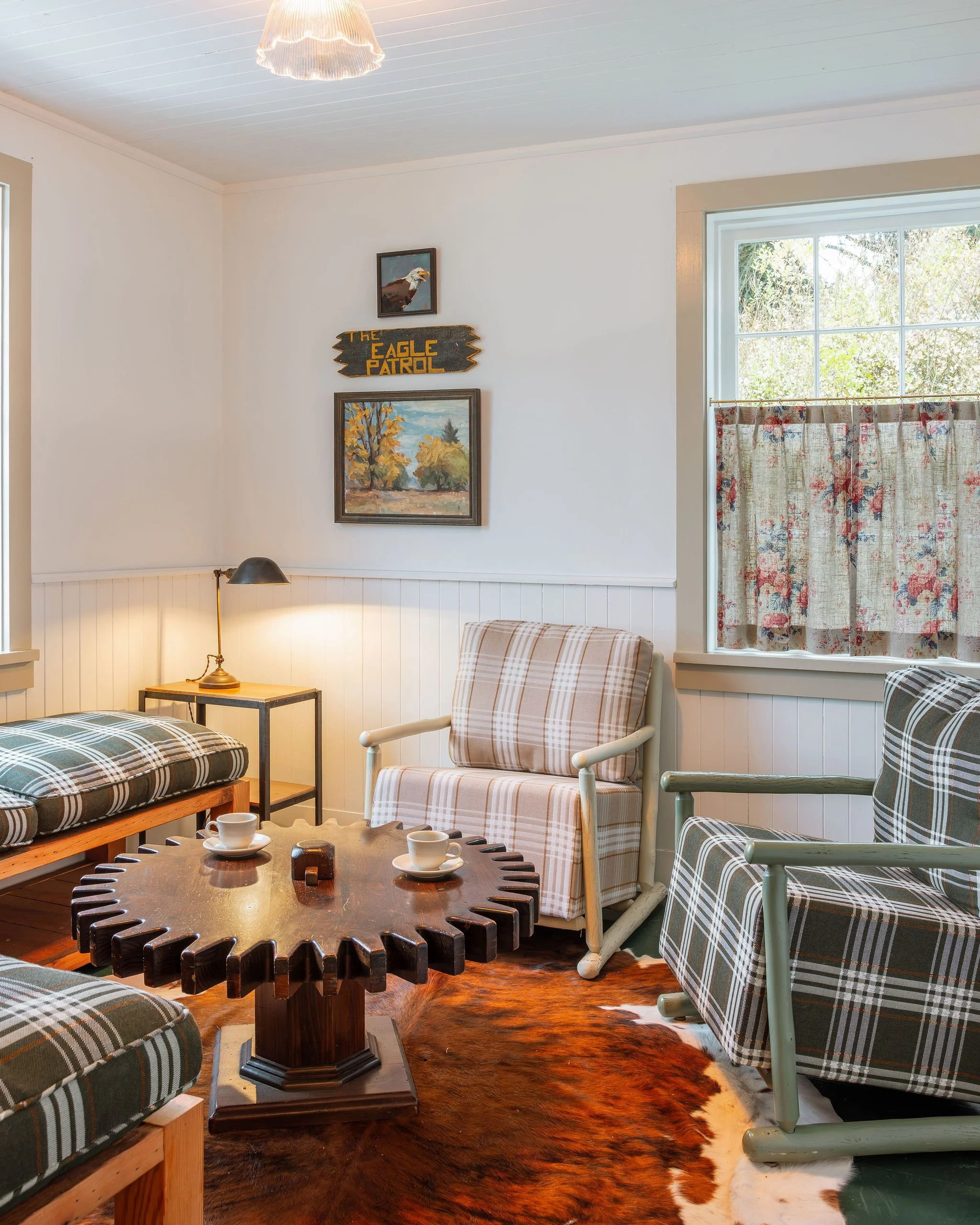 Cozy living room with plaid armchairs, a wooden coffee table with cups, a cowhide rug, and wall decorations including a painting, a 'The Eagle Patrol' sign, and a framed picture, lit by natural light through windows and a hanging ceiling light.
