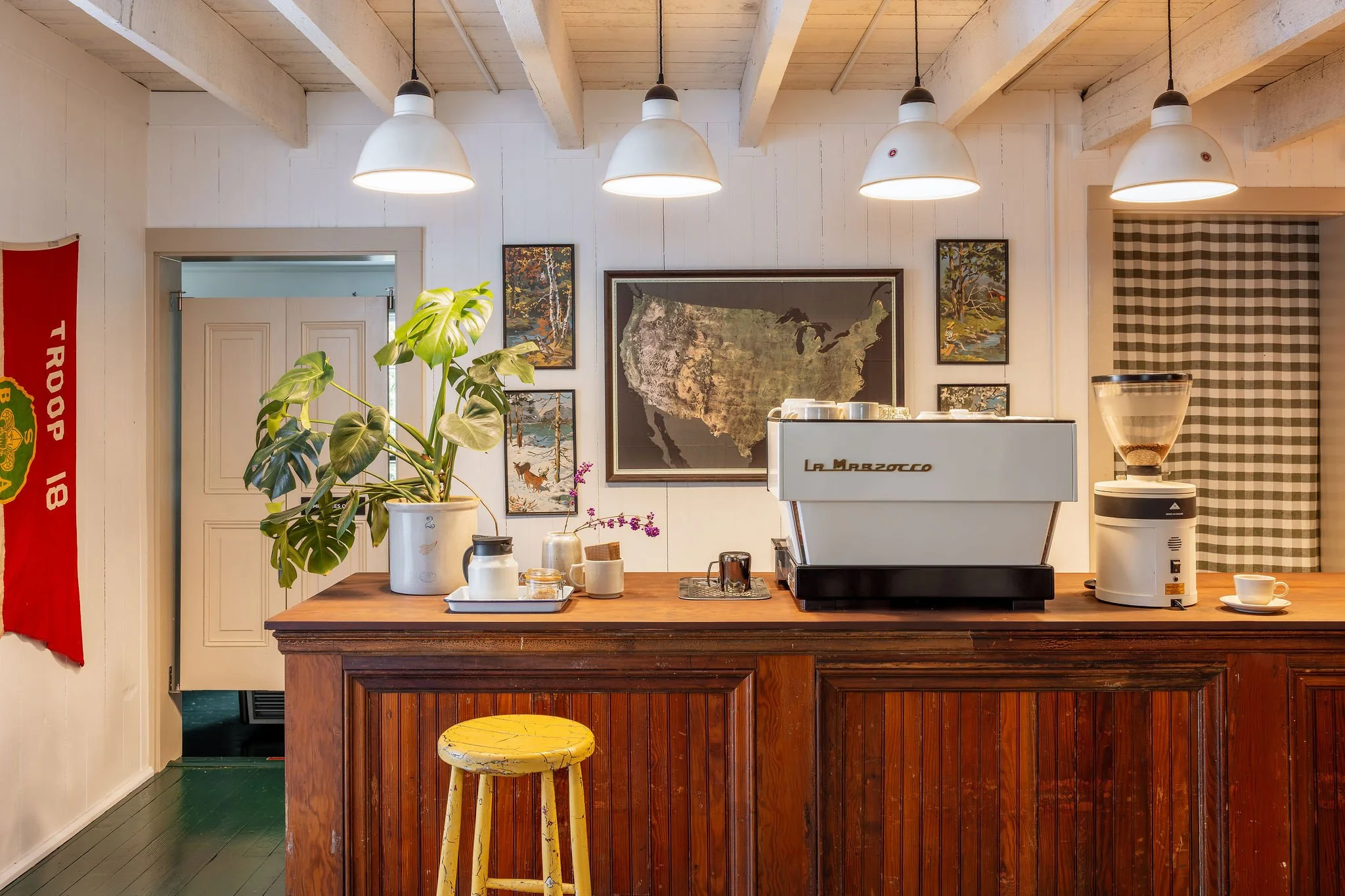 A cozy coffee shop interior with a wooden counter, coffee machine, grinder, green plant, and artwork on white paneled walls.
