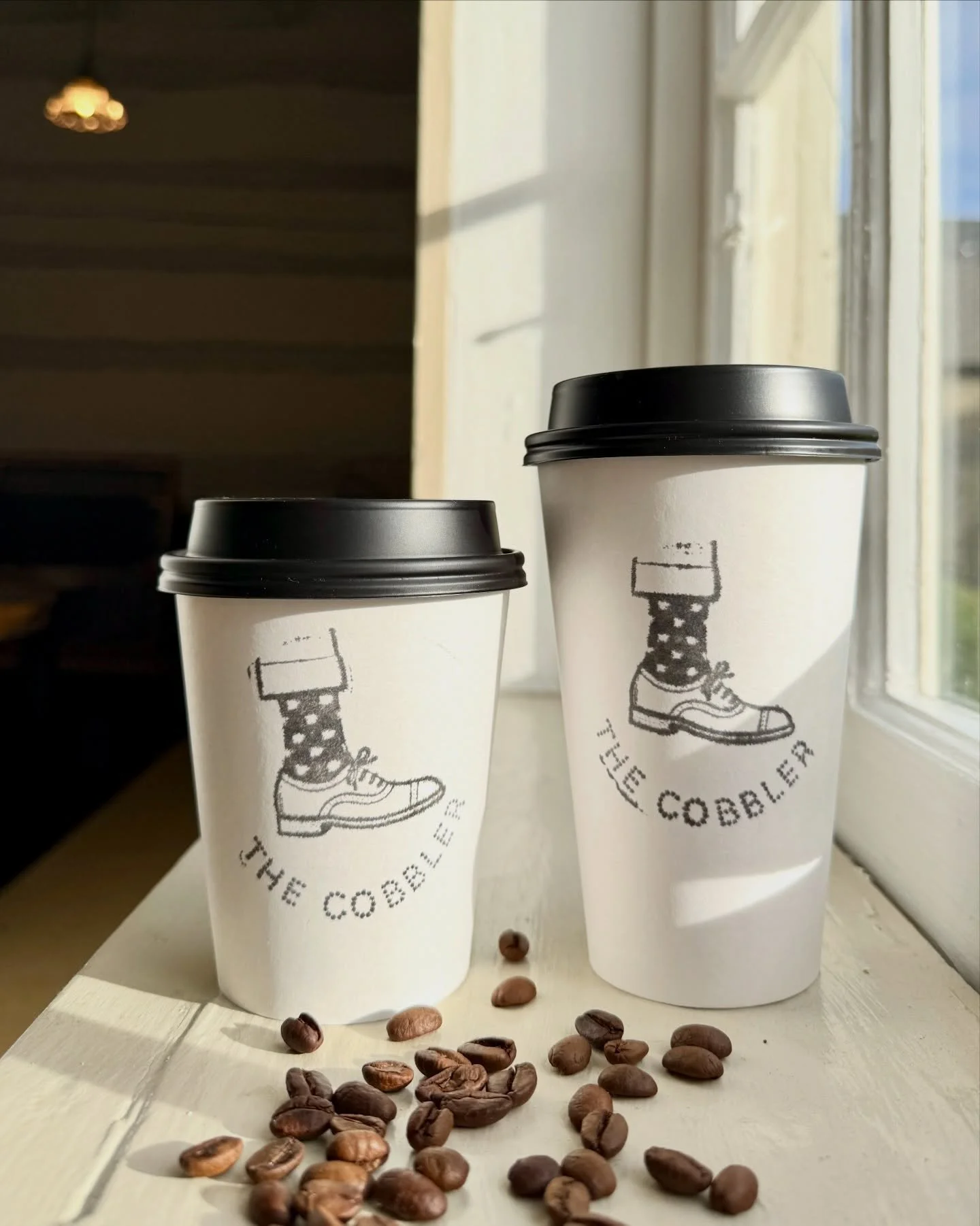 Two white coffee cups with black lids placed on a windowsill, decorated with a drawing of a foot wearing a sock and sneaker, and the words 'THE COBBLER' written underneath. Coffee beans are scattered on the surface in front of the cups, with sunlight streaming through the window.