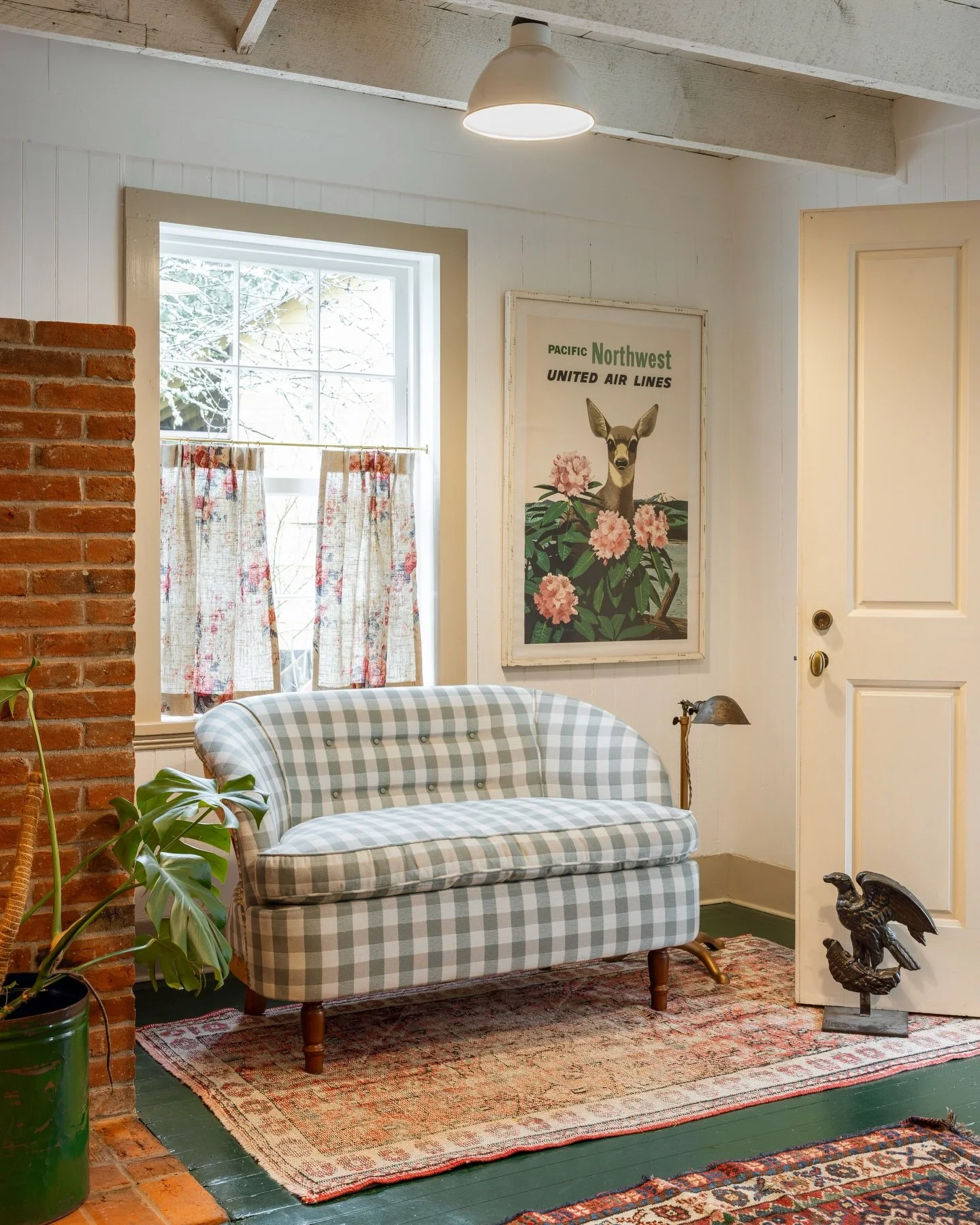 Vintage living room with checkered sofa, potted plant, framed artwork, decorative bird sculptures, window with floral curtains, brick wall, and patterned area rugs.