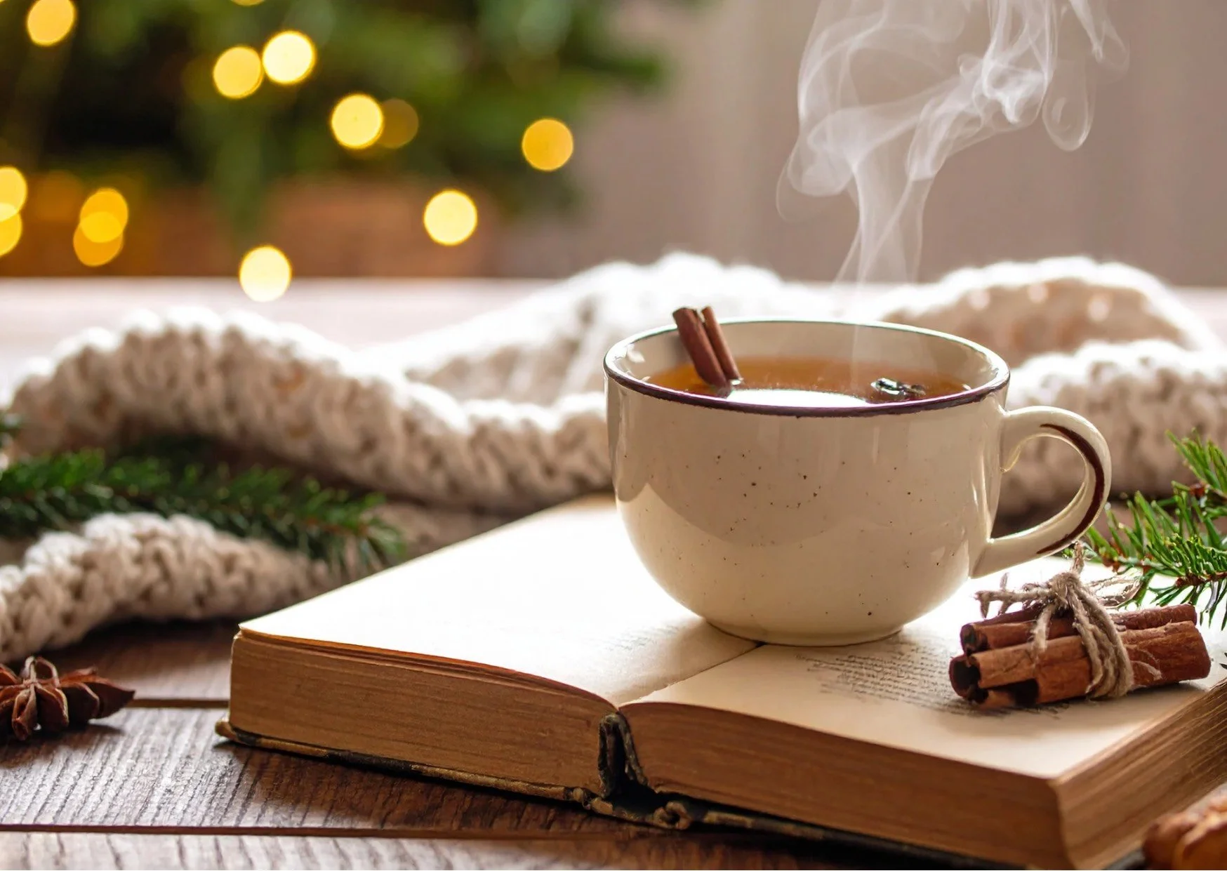 A steamy cup of cinnamon tea sat on an open book, with a bundle of cinnamon sticks placed next to the cup, on the book. A cosy white and green blanket sits behind and a Christmas tree sparkles faintly in the blurred background