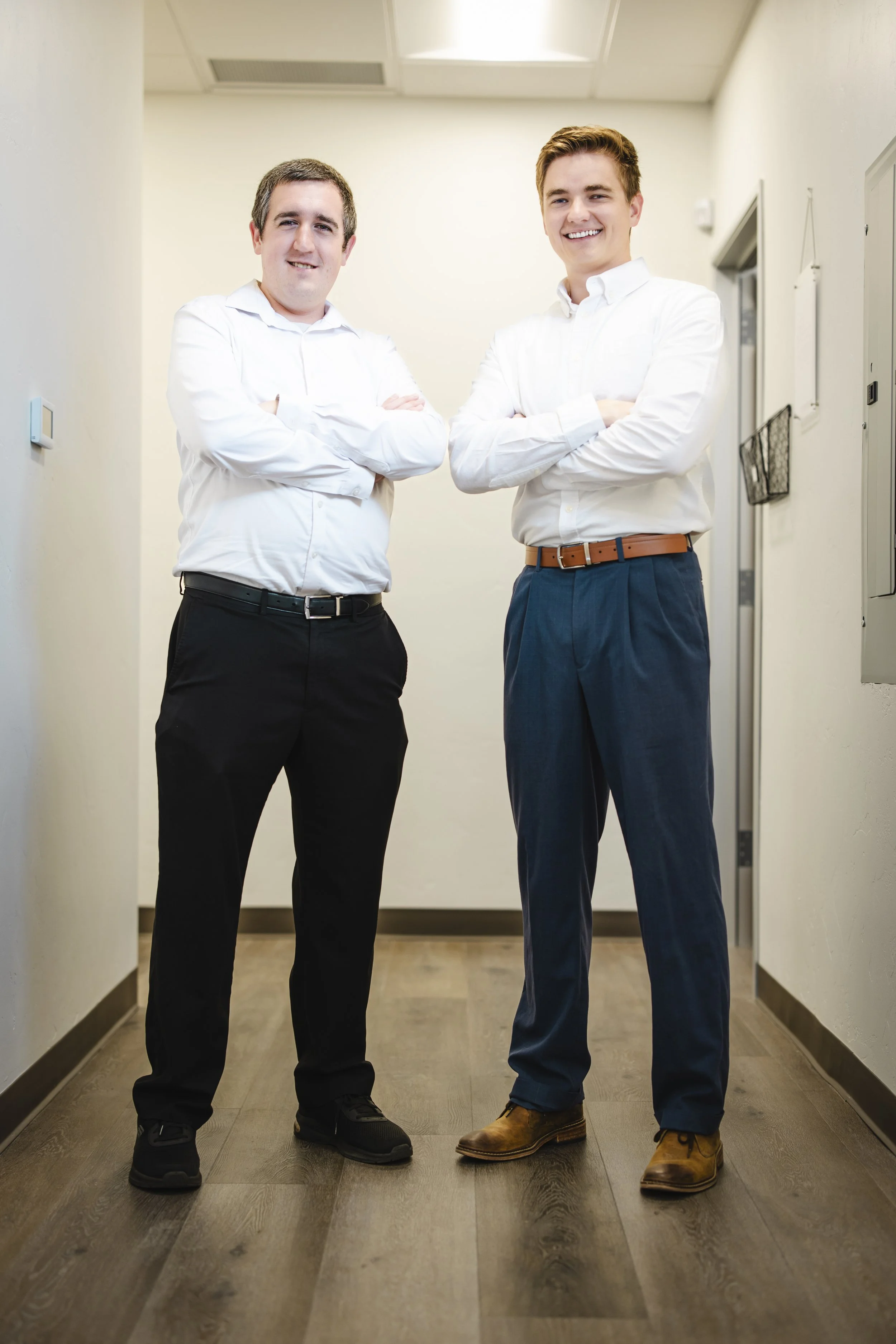 Two young men standing with arms crossed in a hallway, wearing white dress shirts, dark trousers, and dress shoes, smiling at the camera.