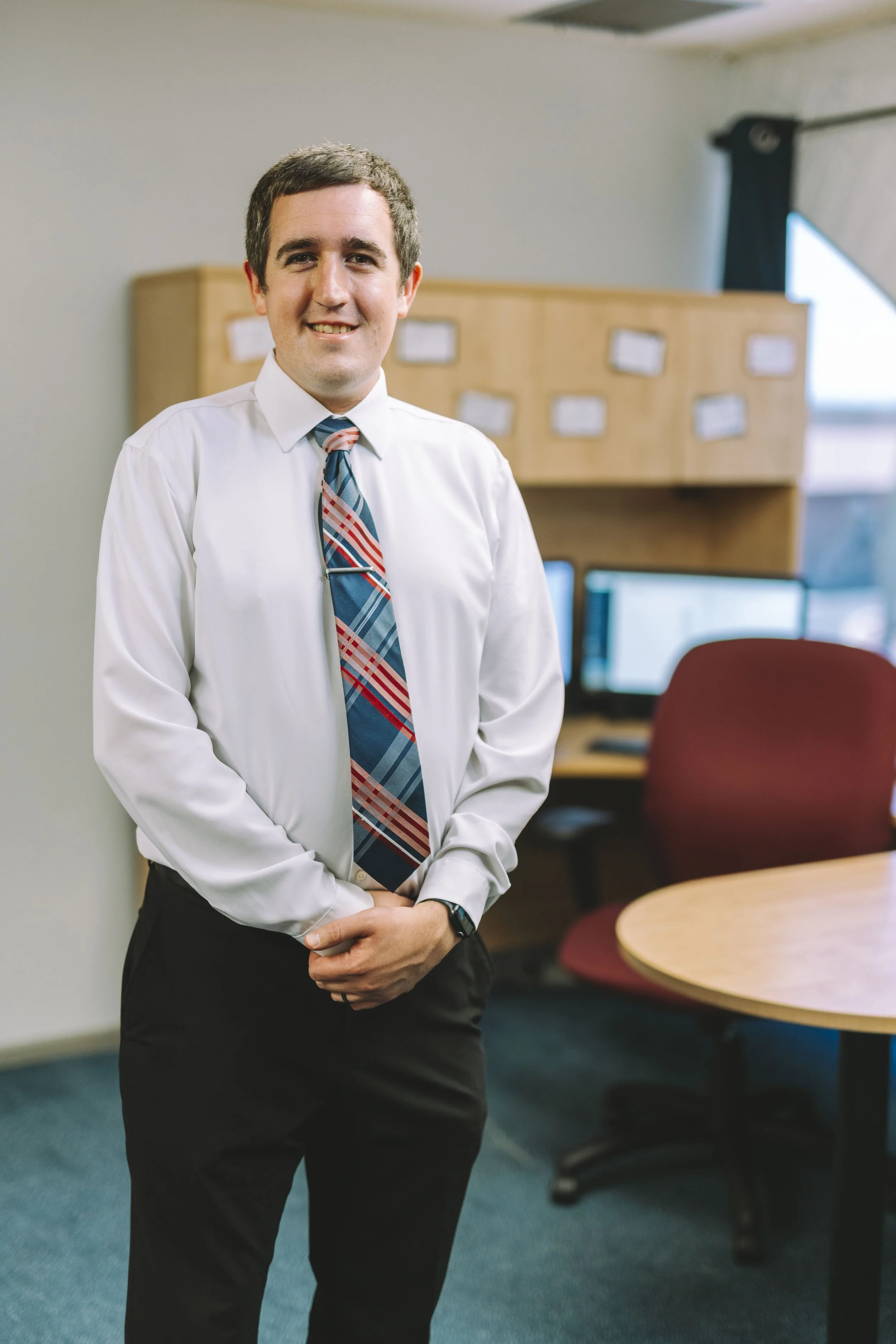 A man in a white dress shirt and a tie standing in an office. There are desks and chairs in the background.