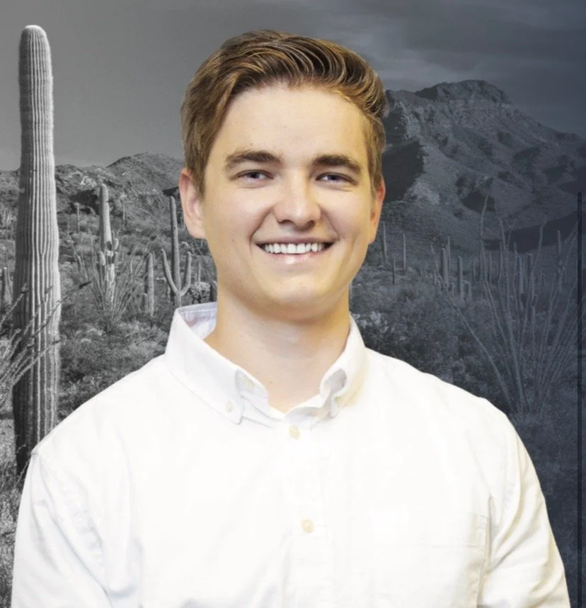 Young man with light brown hair, smiling, wearing a white button-up shirt, standing outdoors with desert mountains and cacti in the background.