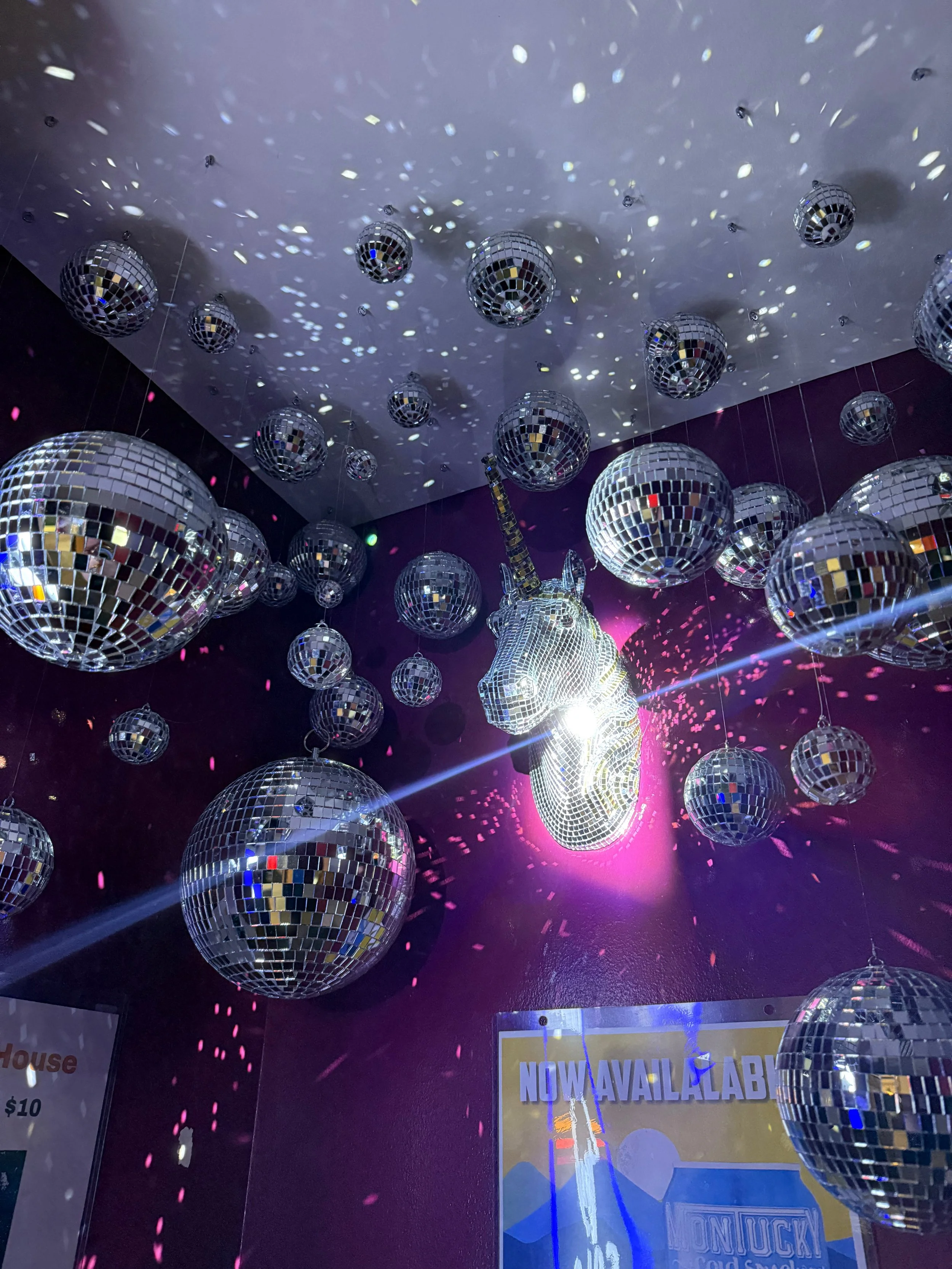 Decorative display of multiple disco balls of various sizes hanging from the ceiling, with the largest disco ball in the shape of a unicorn's head, reflecting light and casting colorful spots on a purple wall.