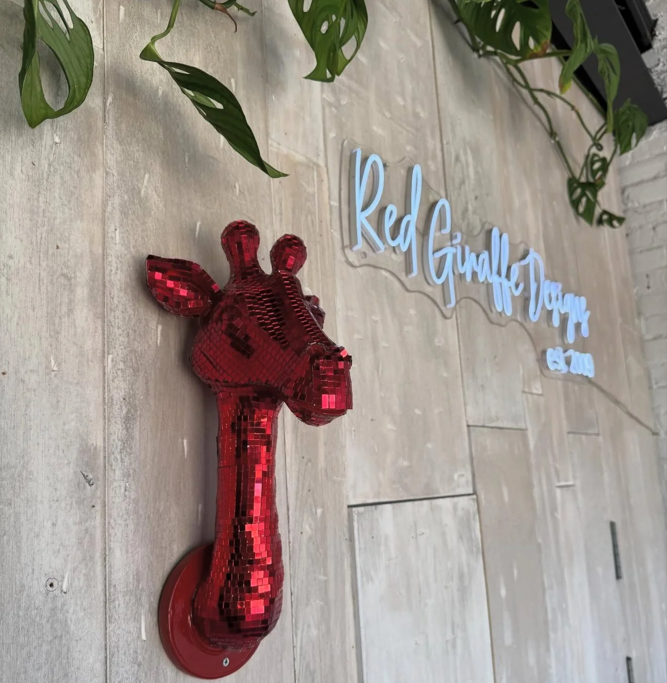 A decorative red giraffe sculpture made of small reflective tiles, placed on a wooden surface with green leafy plants and a blue cursive sign in the background that reads "Red Giraffe Drinks".