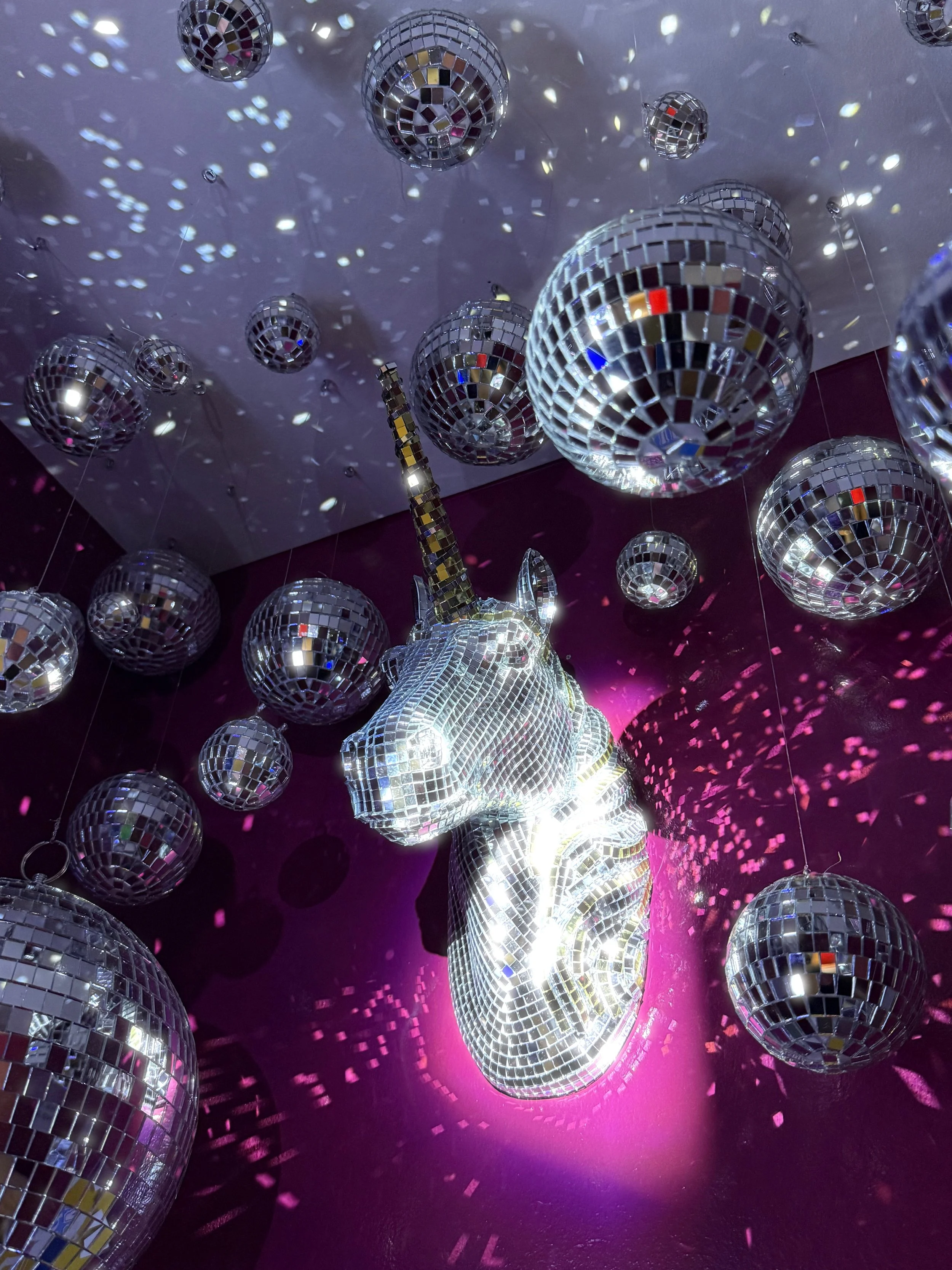 A chandelier shaped like a unicorn head covered in mirror tiles, surrounded by multiple disco balls scattering light reflections in a dark room with pink lighting.