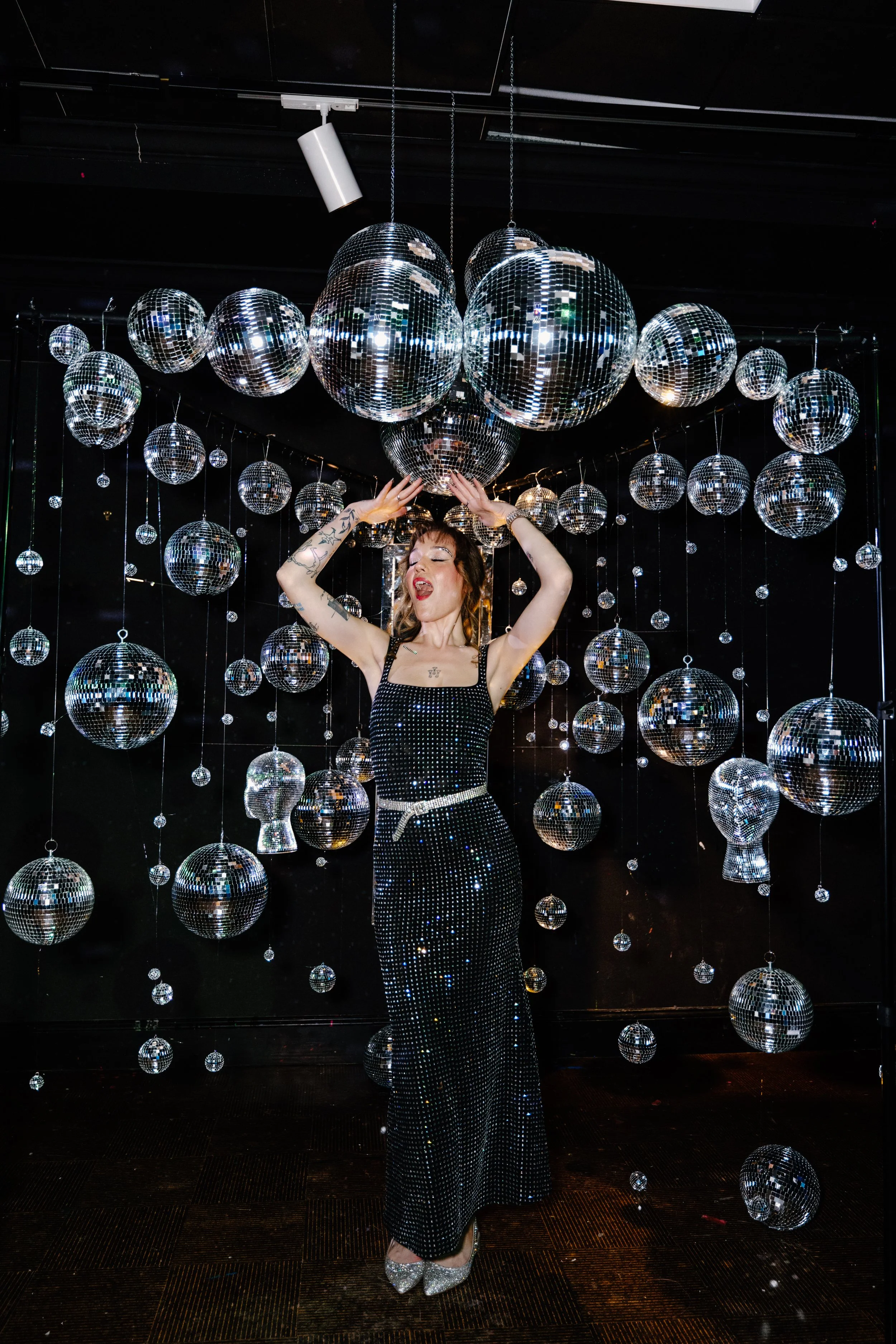 A woman in a sparkly black dress with a white belt is dancing among numerous disco balls of various sizes hanging from the ceiling, creating a lively, festive scene.