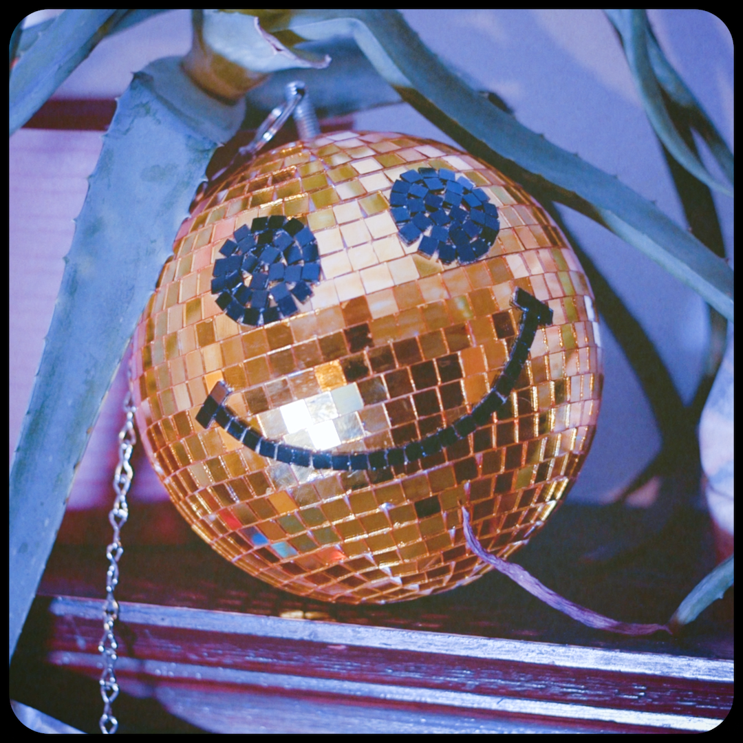 A spherical disco ball with a smiley face design created from small mosaic tiles, hanging among plant leaves.
