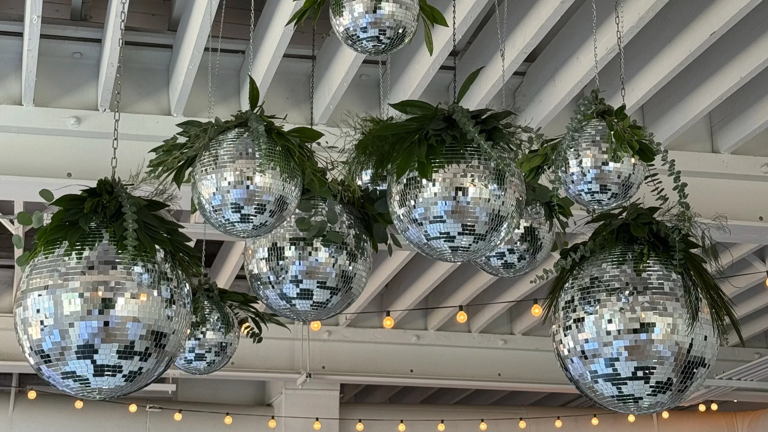 Decorative hanging disco ball ornaments with greenery and plants on a ceiling with string lights.