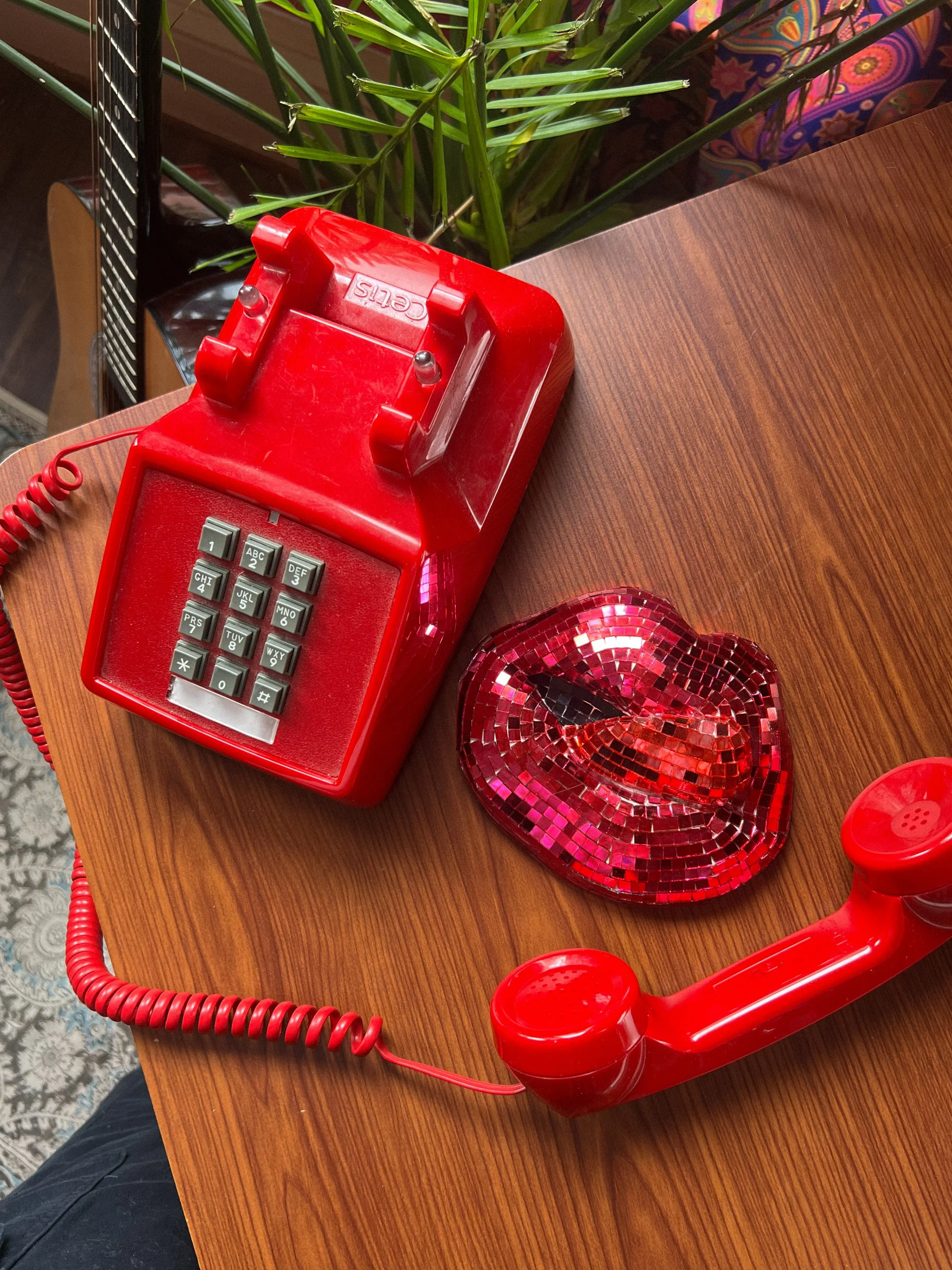 A red vintage rotary phone with a coiled cord, placed on a wooden table. There is a heart-shaped, mosaic, red glass dish nearby and a green plant in the background.
