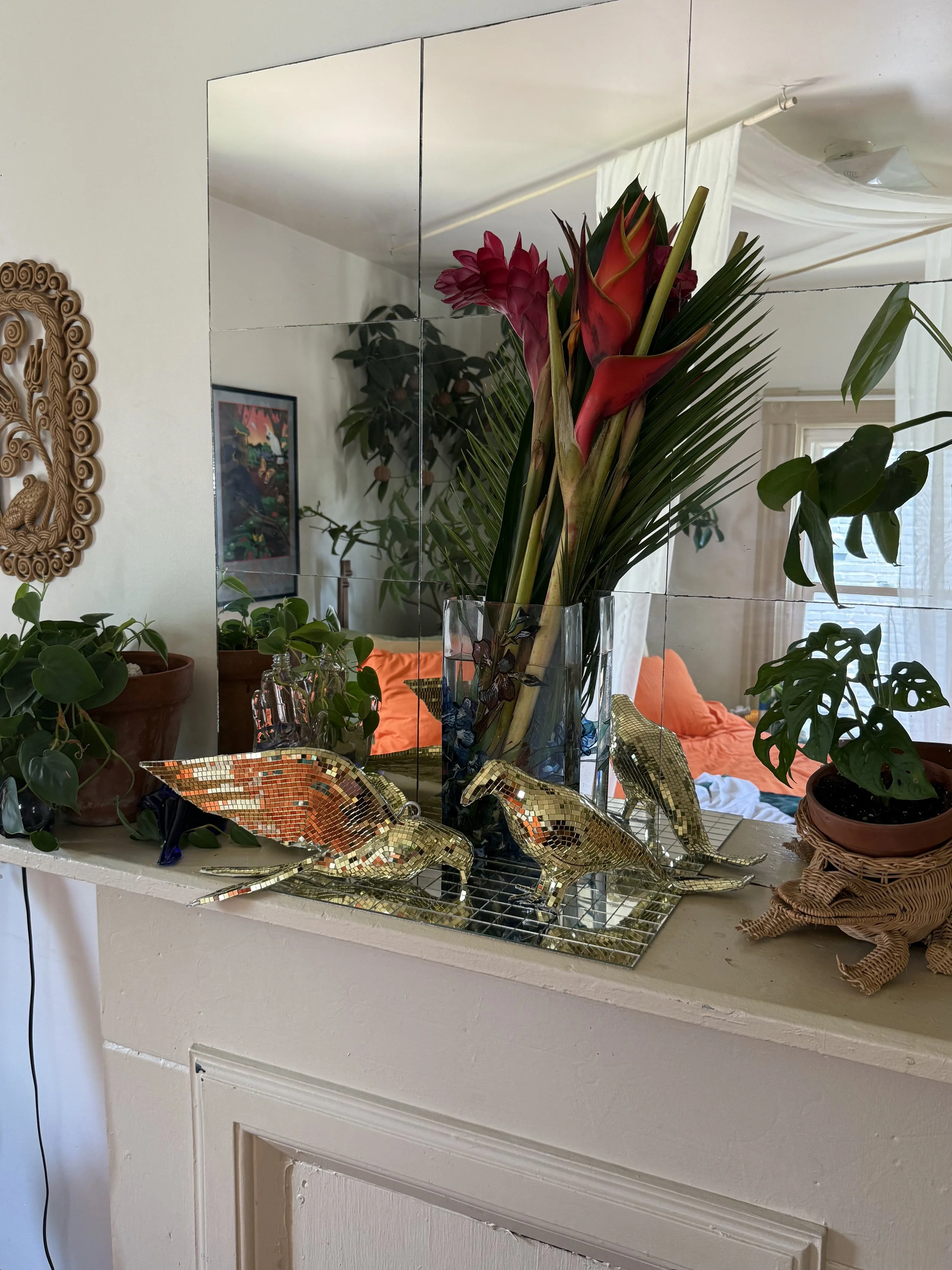 A decorative arrangement on a mantelpiece features a clear glass vase with a bouquet of pink flowers and green foliage, mirrored mosaic animal figurines, and various potted plants. The background shows a living room with a couch, a window with white curtains, and artwork on the wall.