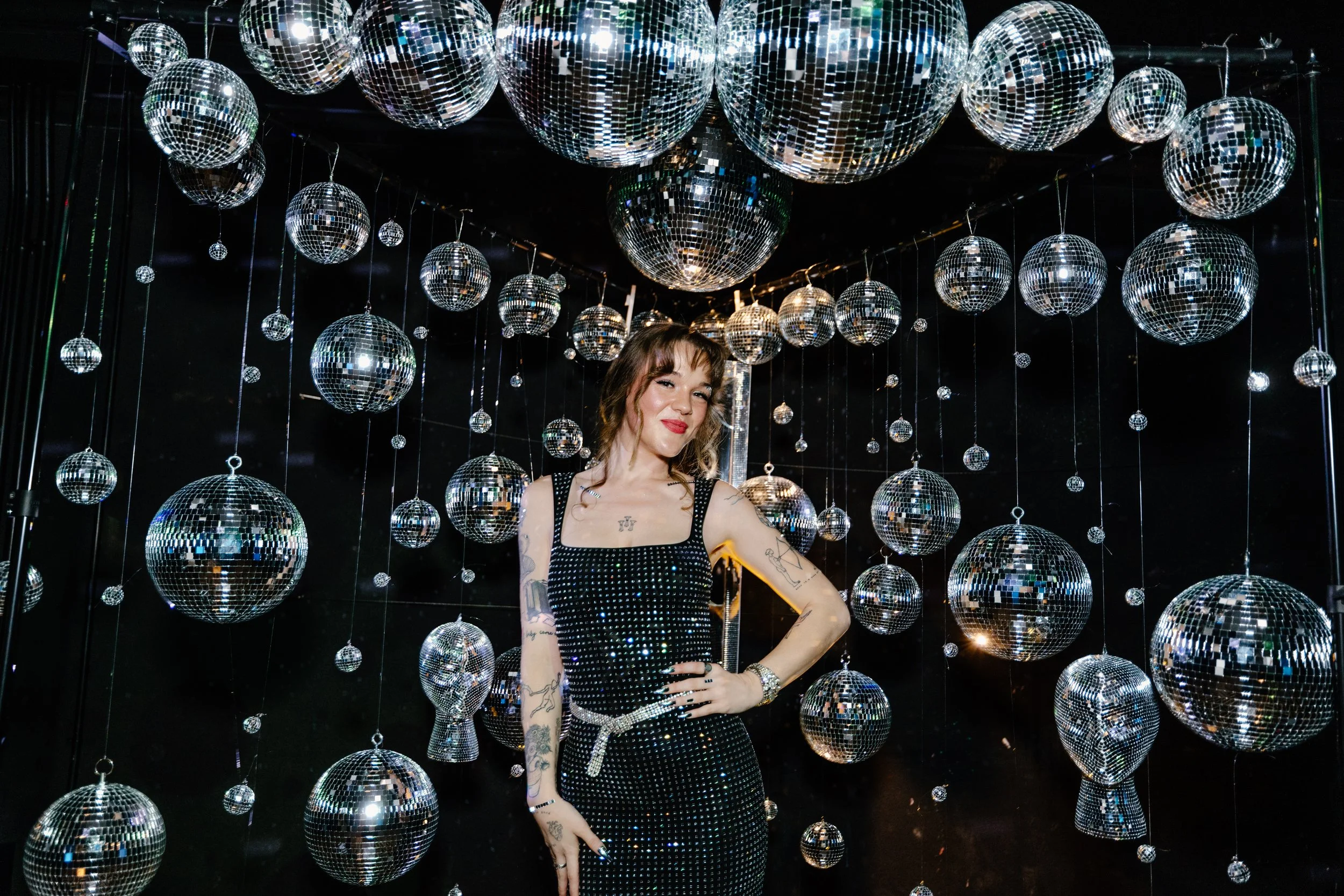 A woman posing in front of numerous hanging disco balls of various sizes in a dark room, wearing a black dress with sparkling embellishments, and smiling at the camera.