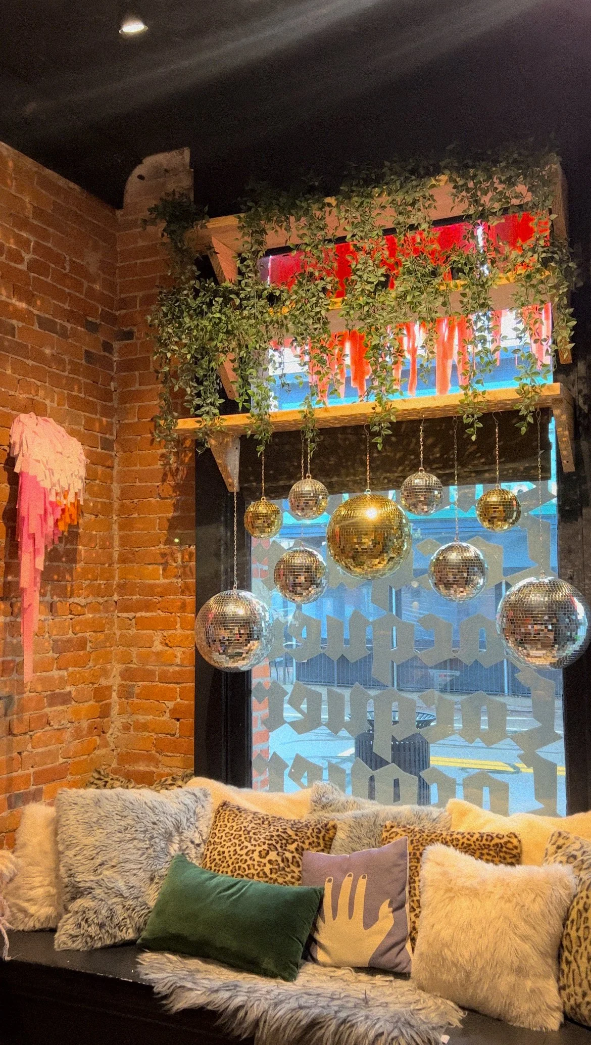 Decorative interior with exposed brick wall, plush pillows, and hanging disco balls near a window with frosted lettering.