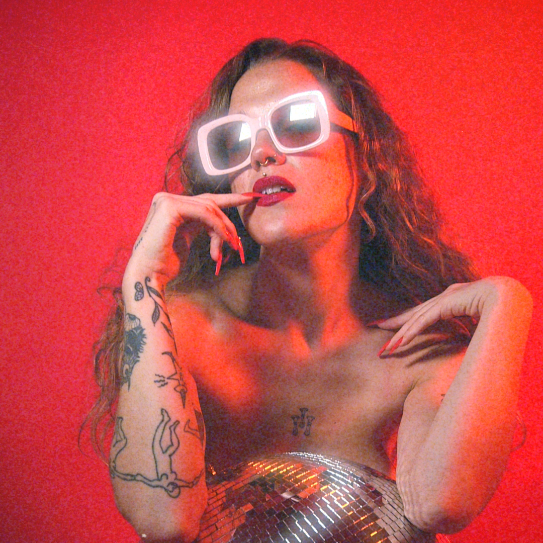 A woman with curly hair wearing large white sunglasses, red lipstick, and tattoos on her arm, posing against a red background with a metallic top.