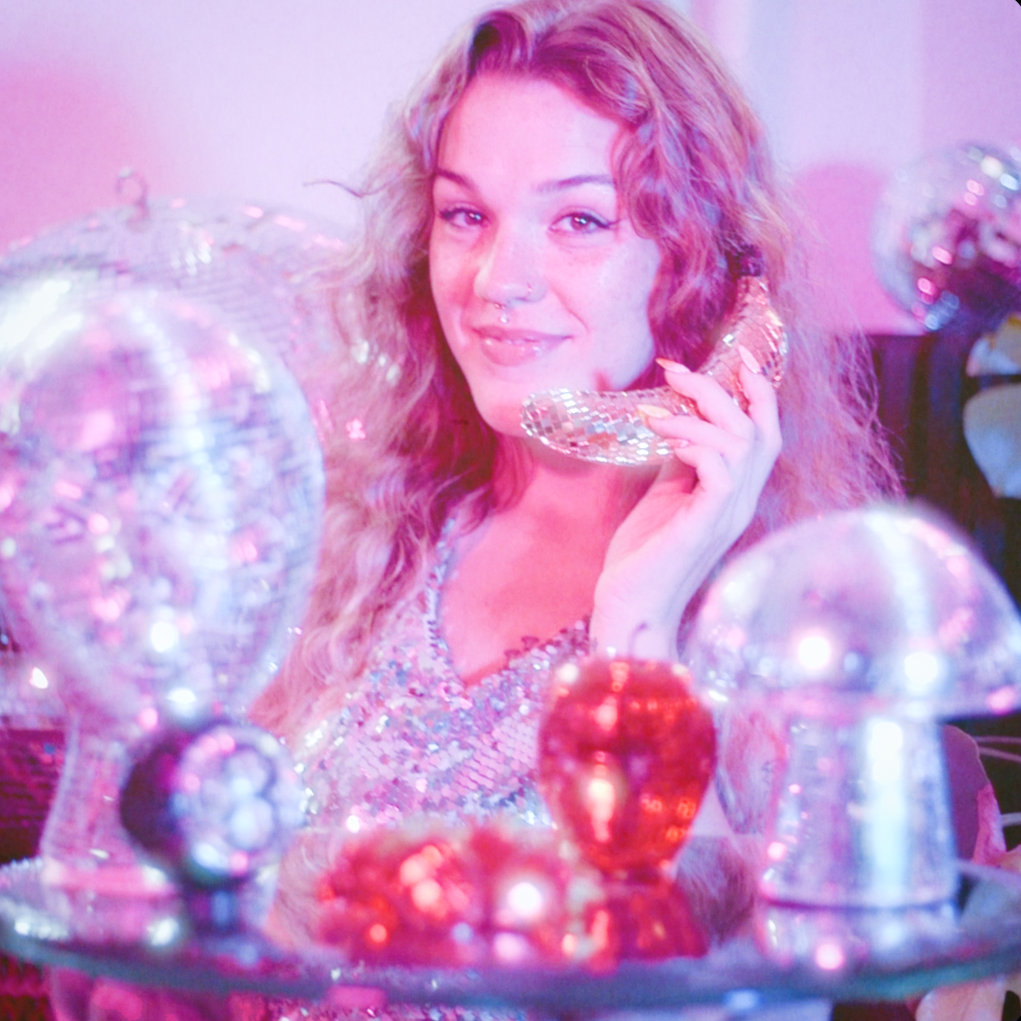 A woman with wavy, light-colored hair and a nose piercing holds a small disco ball while surrounded by various larger disco balls in a room with pink lighting.