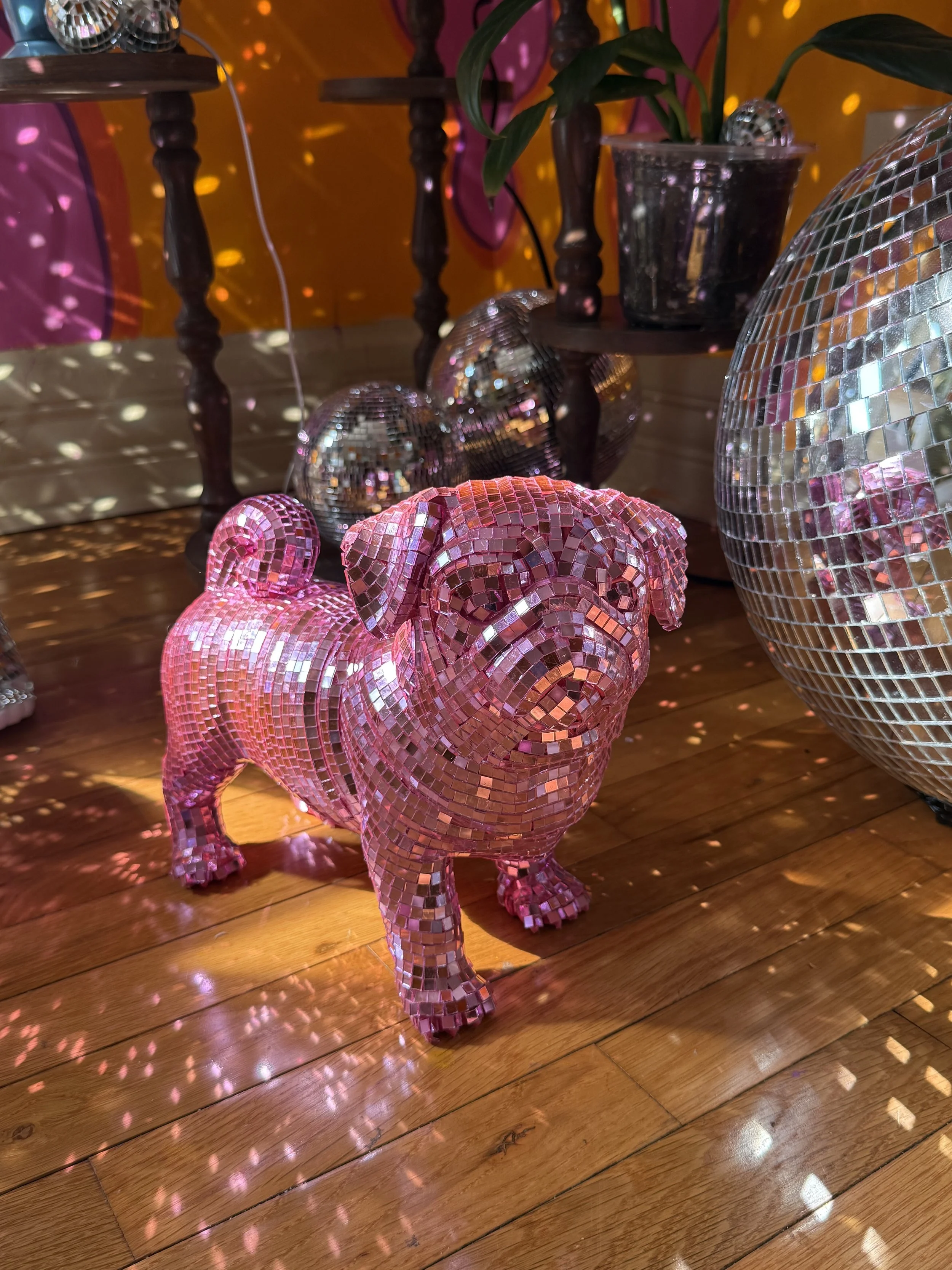 Pink mosaic dog sculpture surrounded by disco balls on a wooden floor with colorful lighting effects.