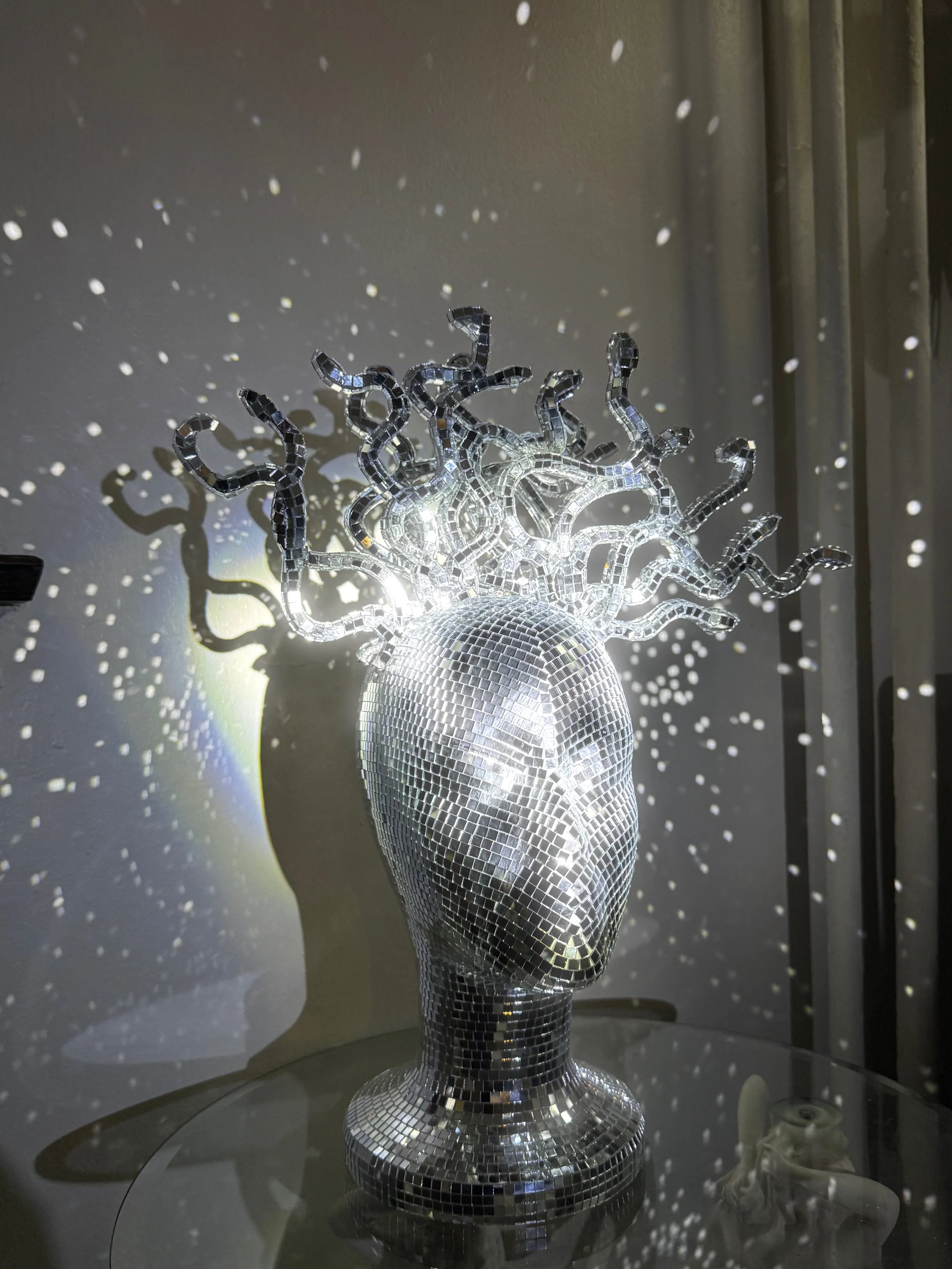 A metallic sculpture of a human head covered in small mirror tiles, with hair made of twisted, reflective, snake-like shapes. The sculpture is illuminated, casting star-like reflections and shadows on the wall behind.