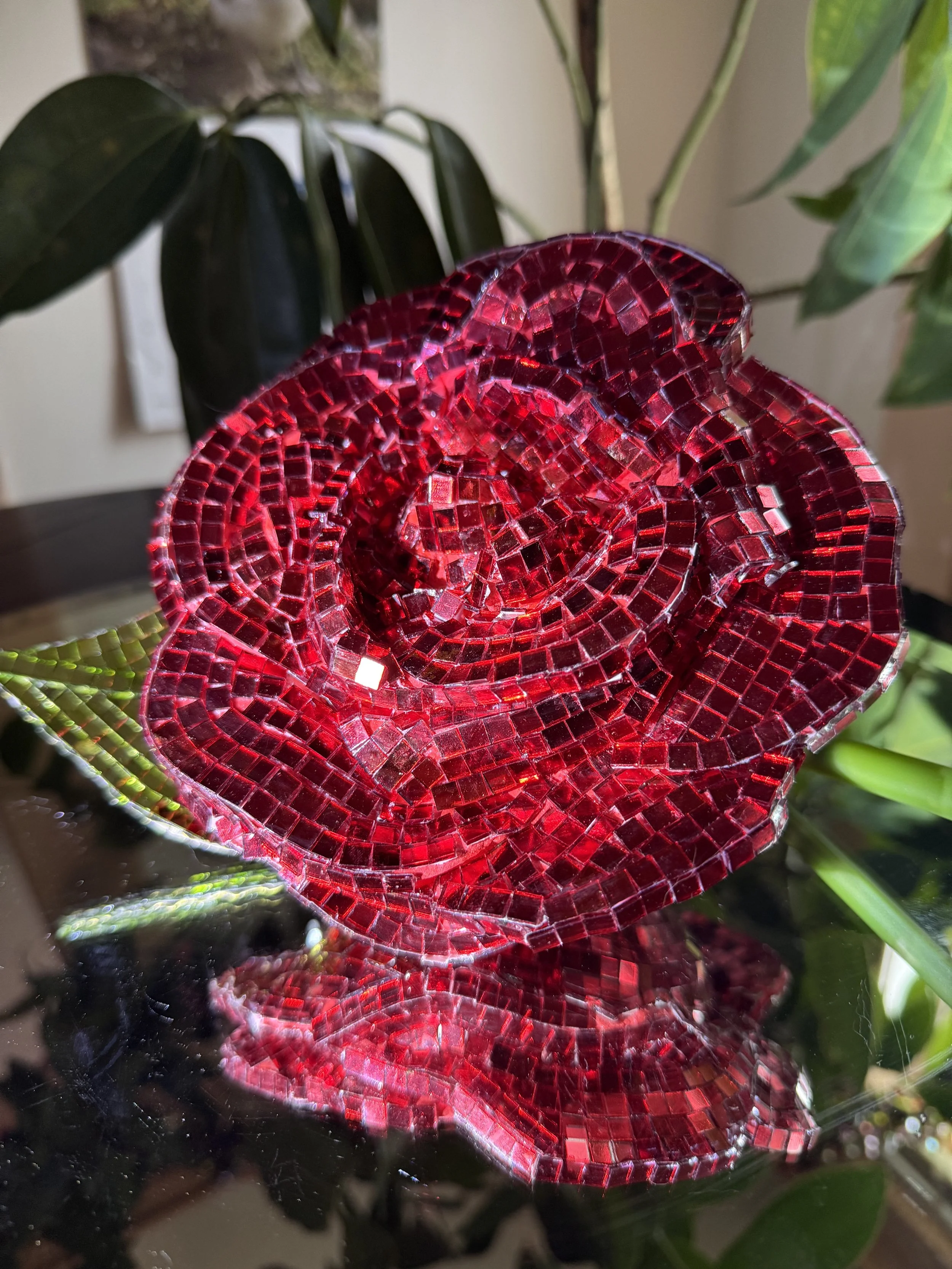 A decorative red mosaic rose sculpture made of small, shiny tiles, placed on a reflective black surface with green plants in the background.