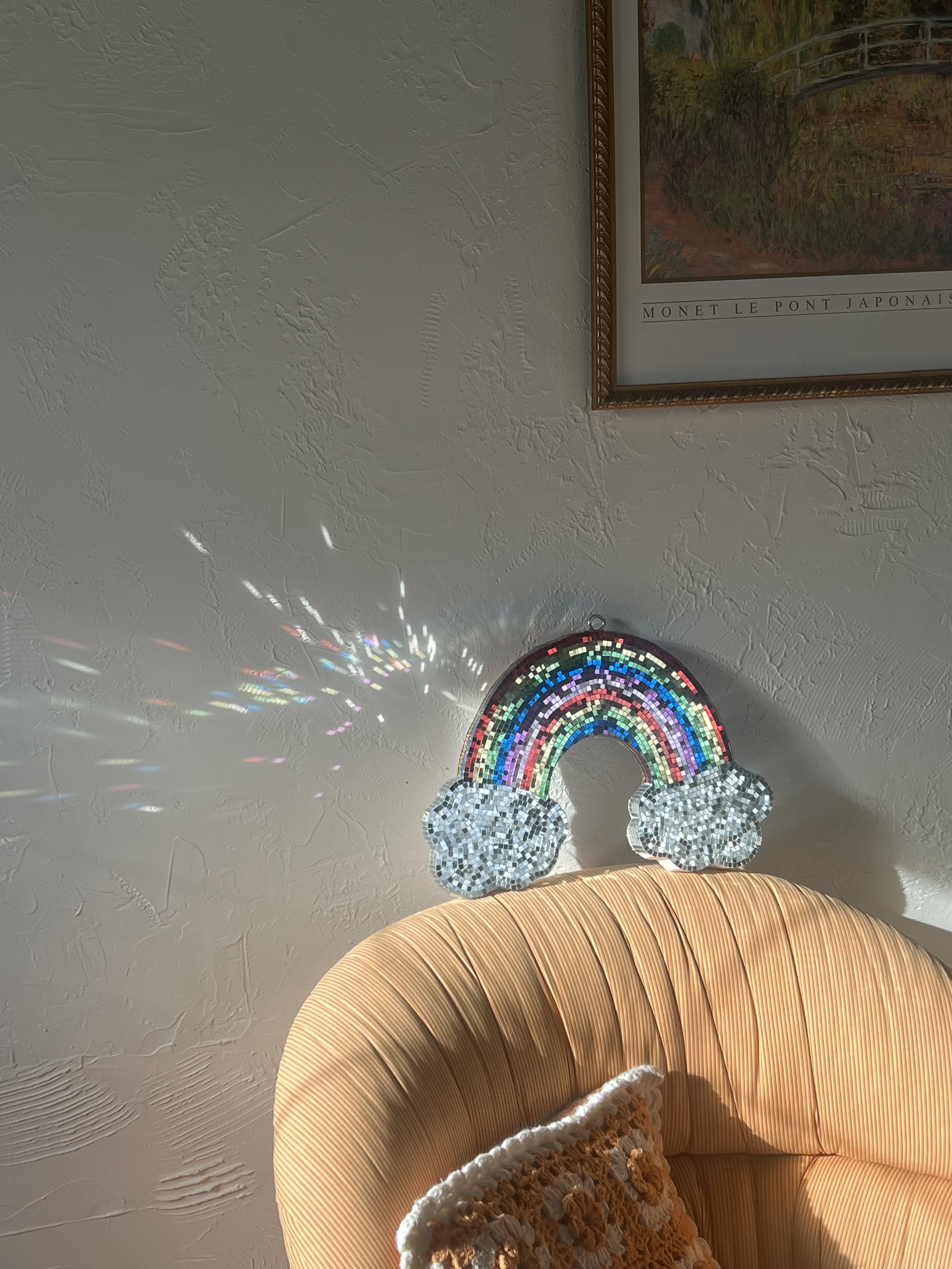 Decorative rainbow wall hanging made of small, colorful tiles and mirrored clouds, casting a rainbow and light reflections on a textured beige wall.