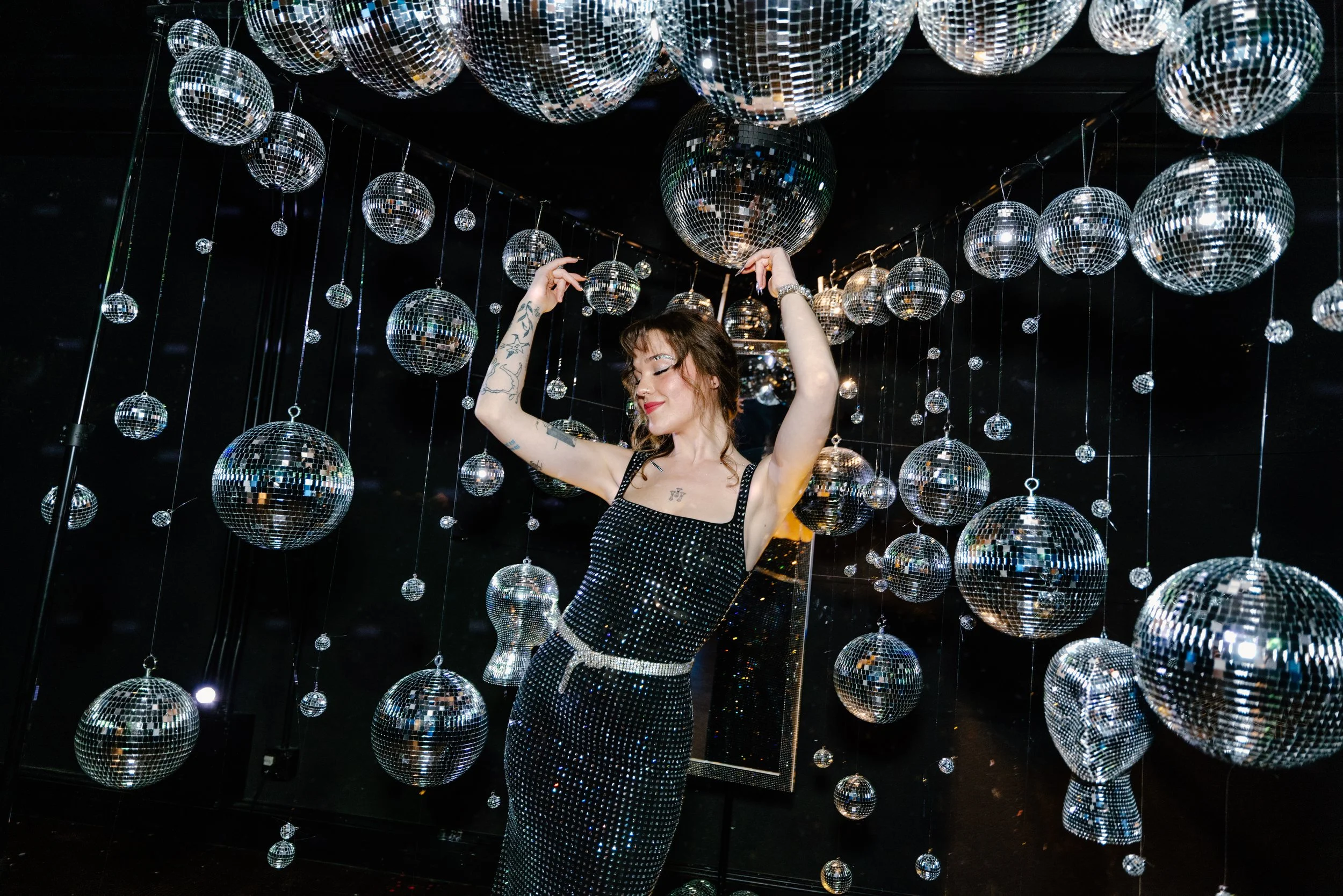 A woman in a black, sequined dress with a white belt is dancing among hanging mirrored disco balls of various sizes in a dark room.