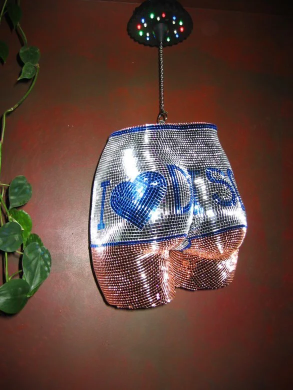 A metallic purse decorated with shiny rhinestones, featuring a design with the words "I love DIS" and a heart symbol, hanging from a ceiling. There is a green plant with large leaves on the left side of the image.