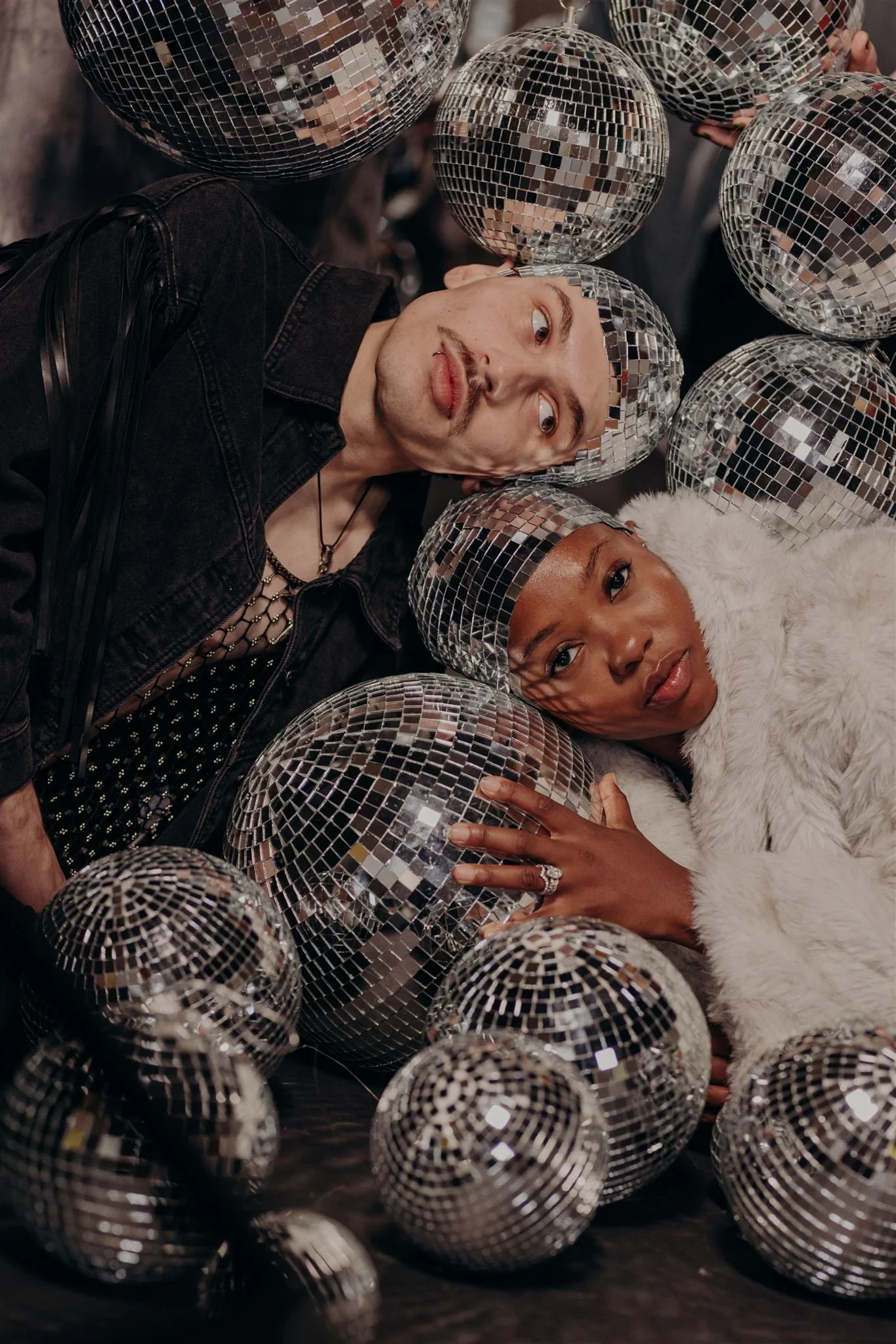 A man and woman lie on the floor among multiple disco balls, surrounded by their reflected images. The man wears a black denim jacket, and the woman has a ring on her finger and wears a white furry coat.