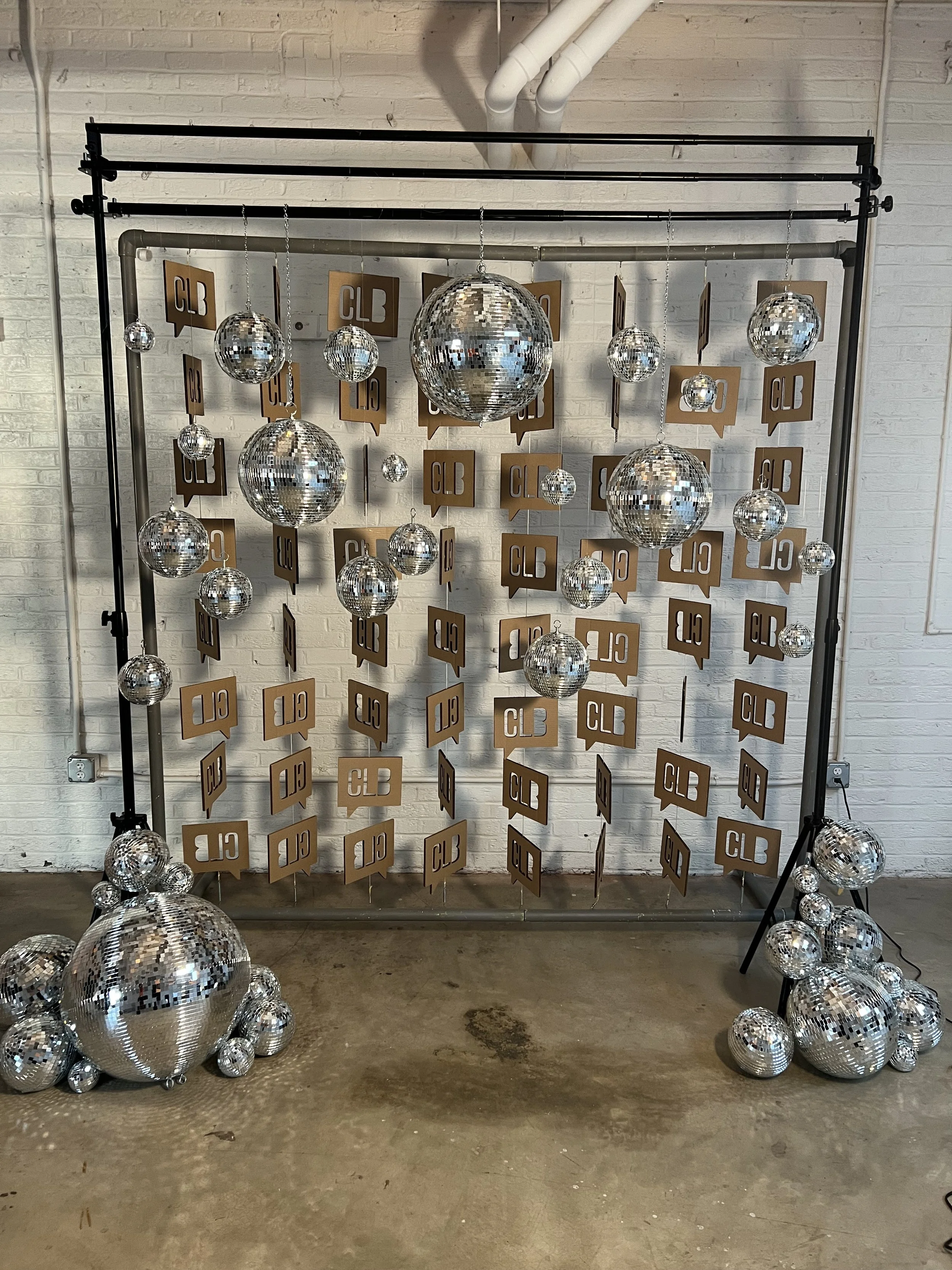 Decorative display with multiple disco balls of various sizes hanging from a metal frame, with additional disco balls on the ground, and gold signs with the letters 'CLB' hung on the background wall.
