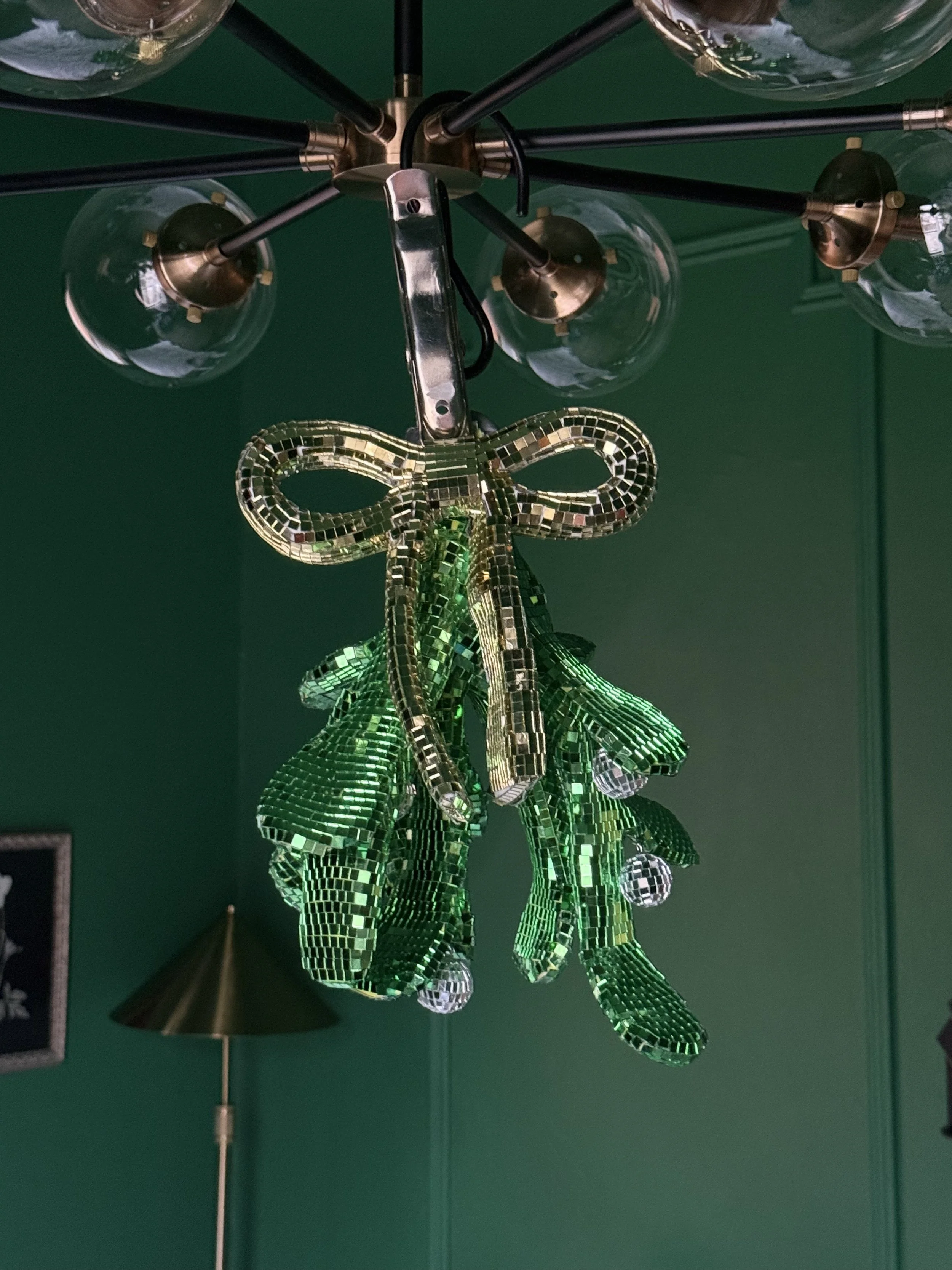 A modern chandelier with multiple glass light bulbs and a decorative hanging piece made of green and silver tiles resembling leaves, hanging from the chandelier's metal fixture, against a dark green wall.