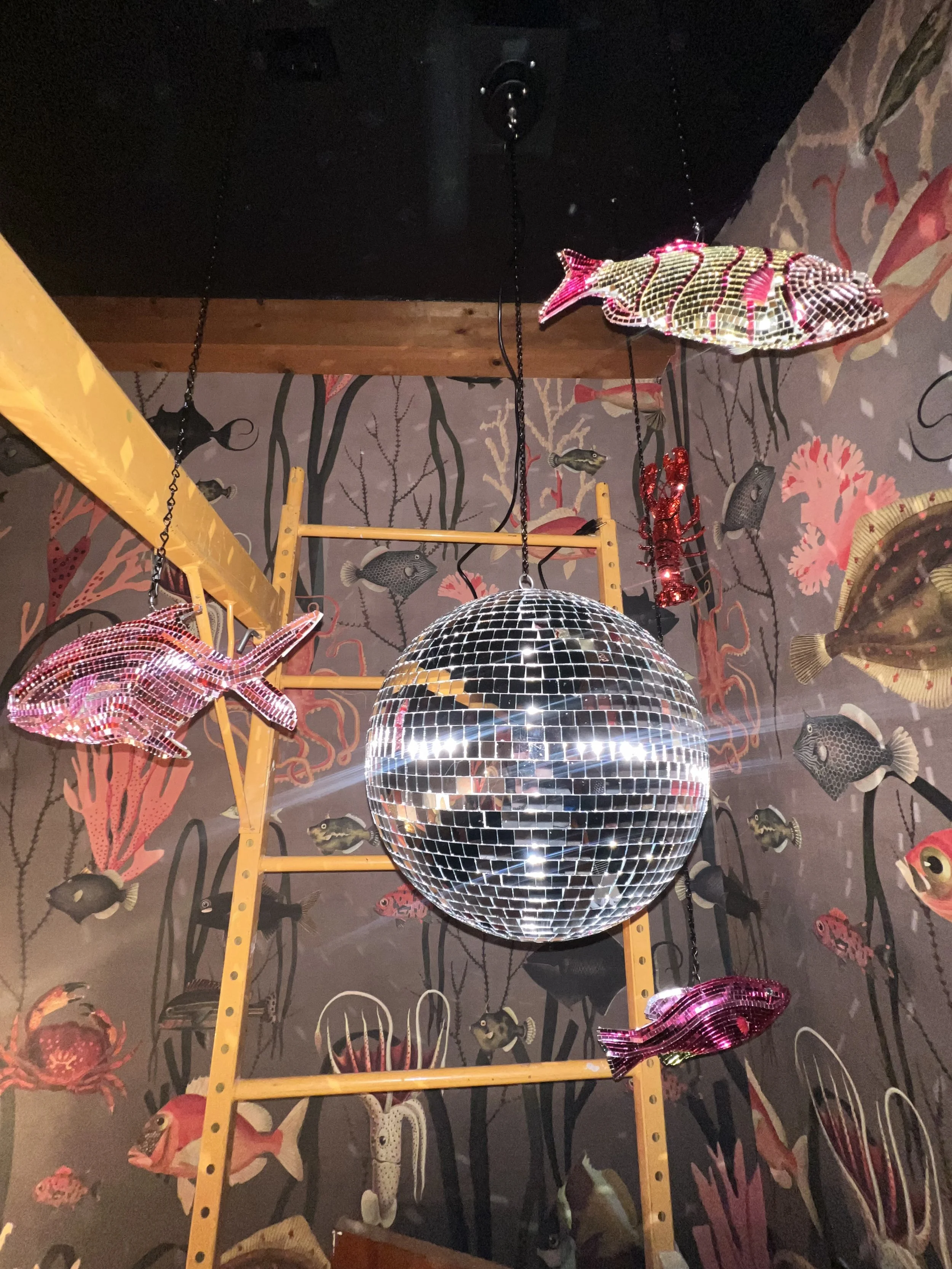 Disco ball chandelier with decorative fish sculptures hanging from the ceiling, in a room with an underwater themed mural.
