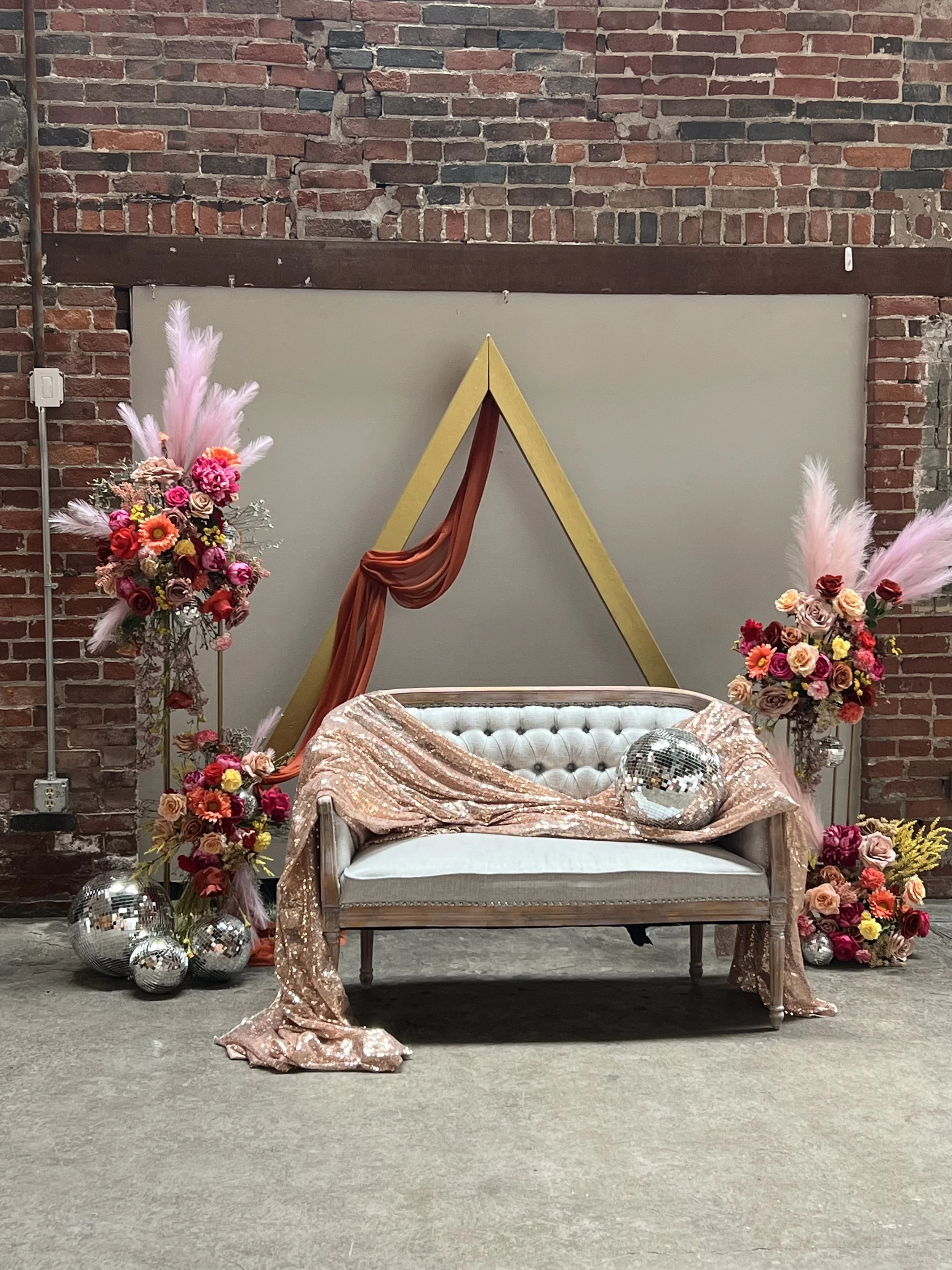 Decorative setup with a vintage-style white upholstered sofa draped with pink sequined fabric, adorned with disco balls, and surrounded by vibrant flower arrangements and pampas grass against an industrial brick wall background.