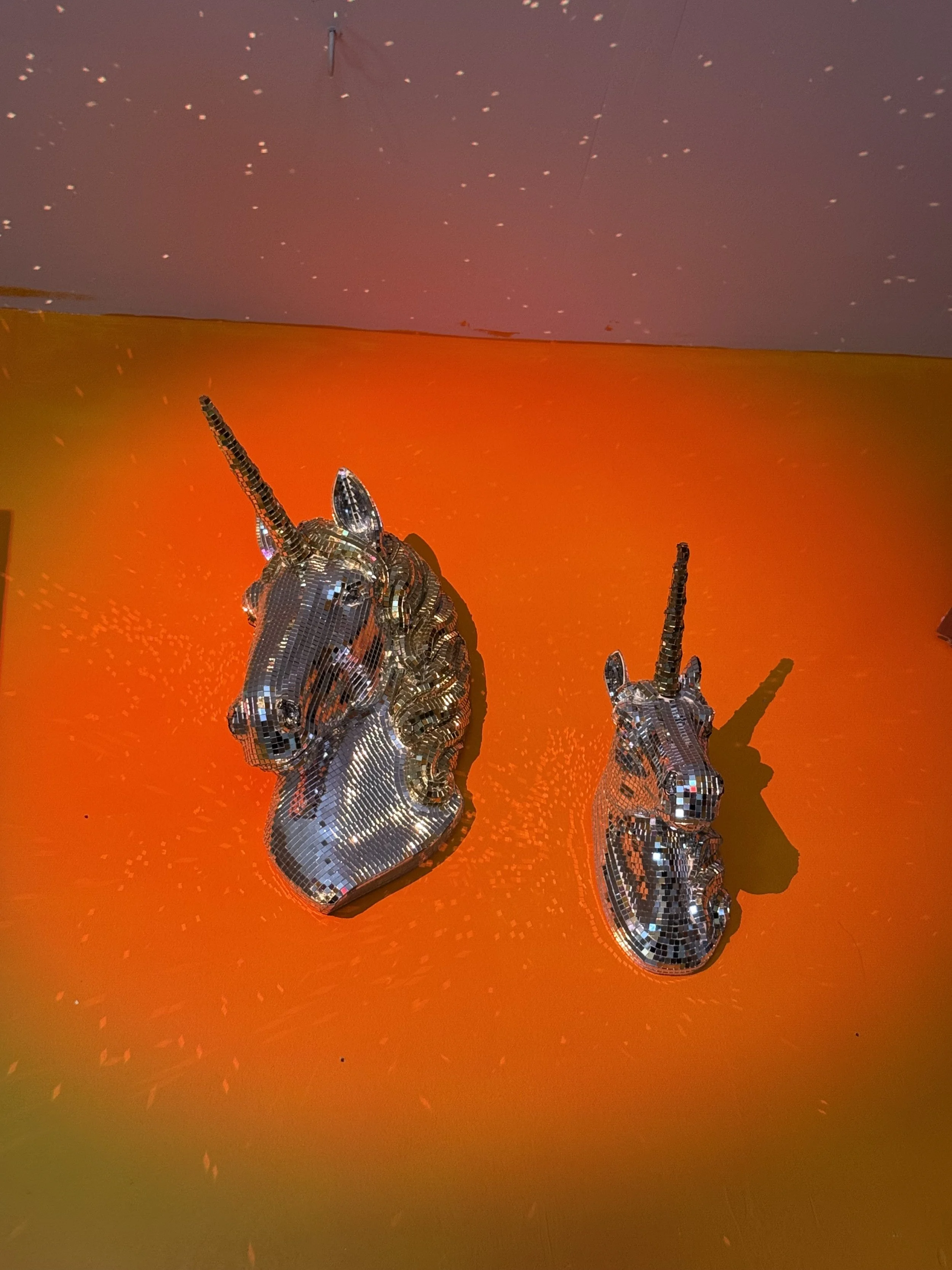 Two metallic unicorn head sculptures with shimmering, mirror-like surfaces on an orange background, casting shadows.