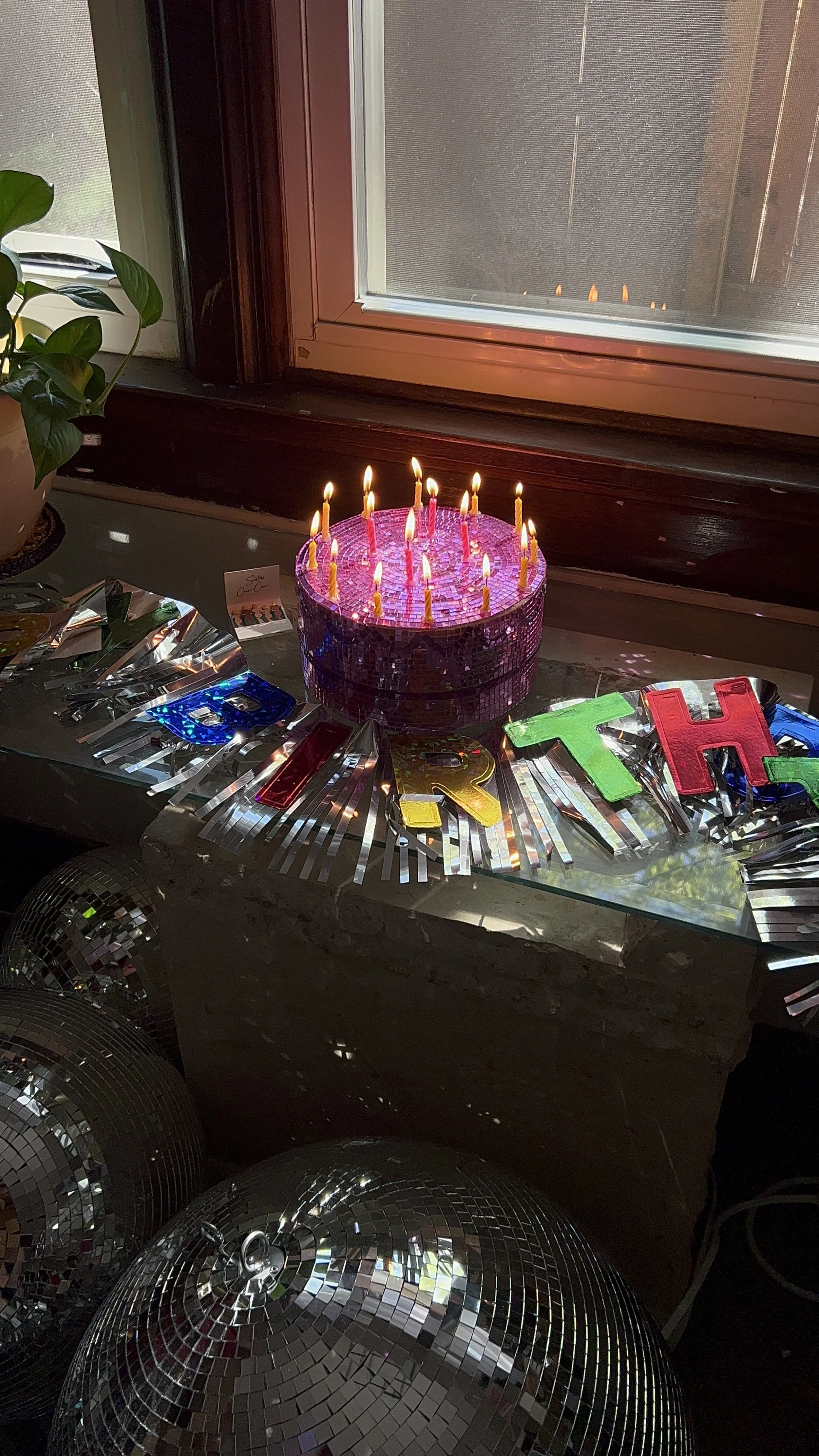 A birthday cake with lit candles on top, surrounded by festive decorations including shiny letter banners spelling 'HAPPY BIRTHDAY' on a glass table, with disco balls underneath and a window in the background.