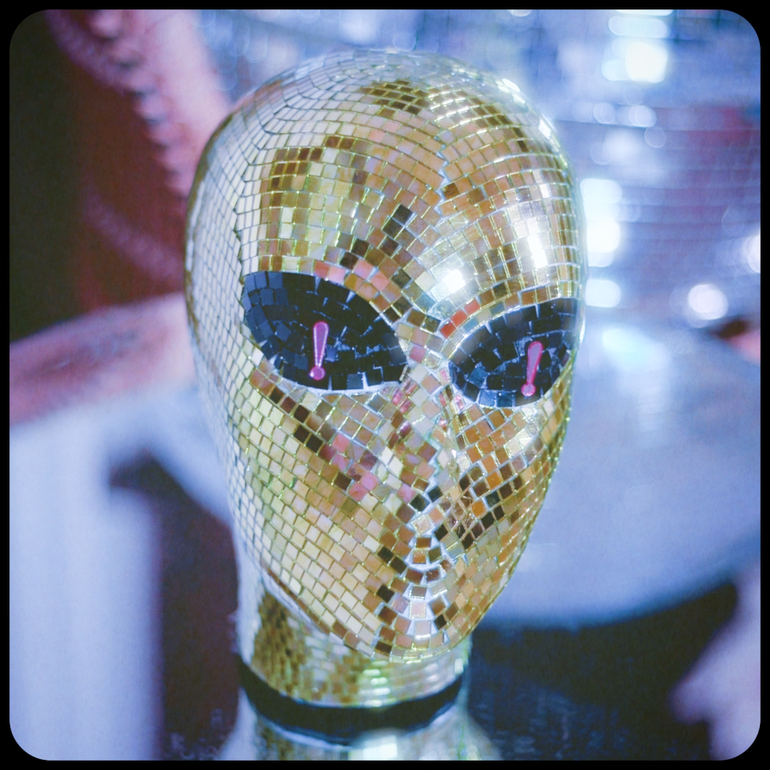 A shiny gold-colored humanoid head sculpture covered in mirrored tiles with two large black eyes featuring pink exclamation marks as pupils, set against a blurred background with bright lights.