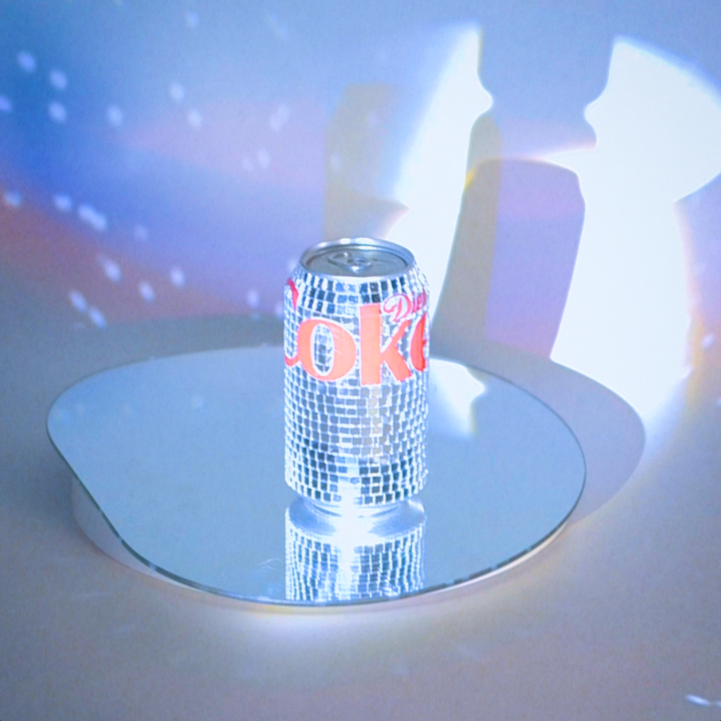 A can with a mosaic tile pattern and red lettering that says "OK" on a reflective round surface, with a colorful light display projecting abstract patterns on the wall behind it.