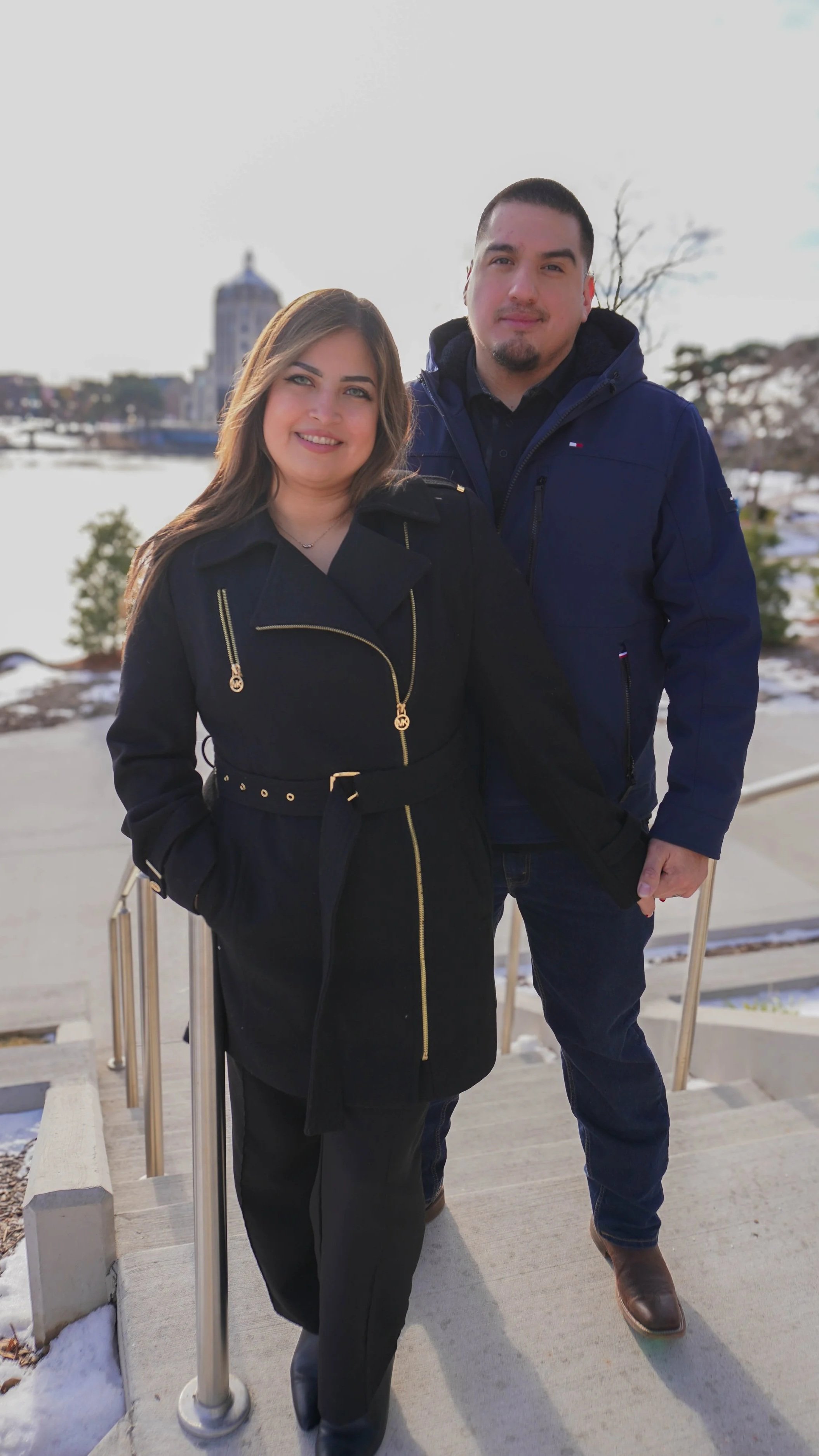 A smiling woman and a man standing outdoors near a river or lake during winter, dressed in dark jackets, with city buildings in the background.