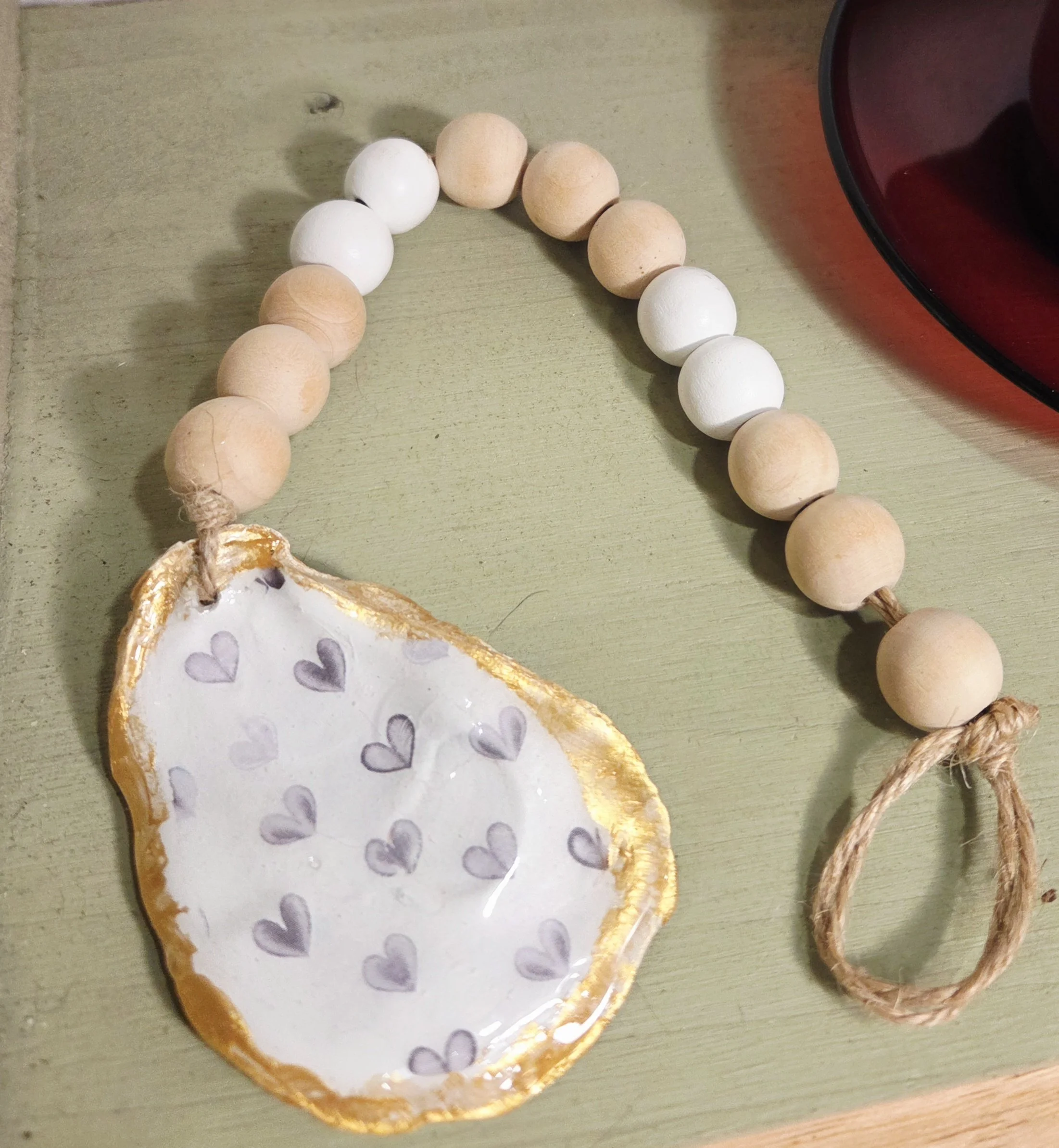 Decorative beaded garland with white and raw wood round beads, painted oyster shell with gray heart designs and gold trim resting on a white surface.