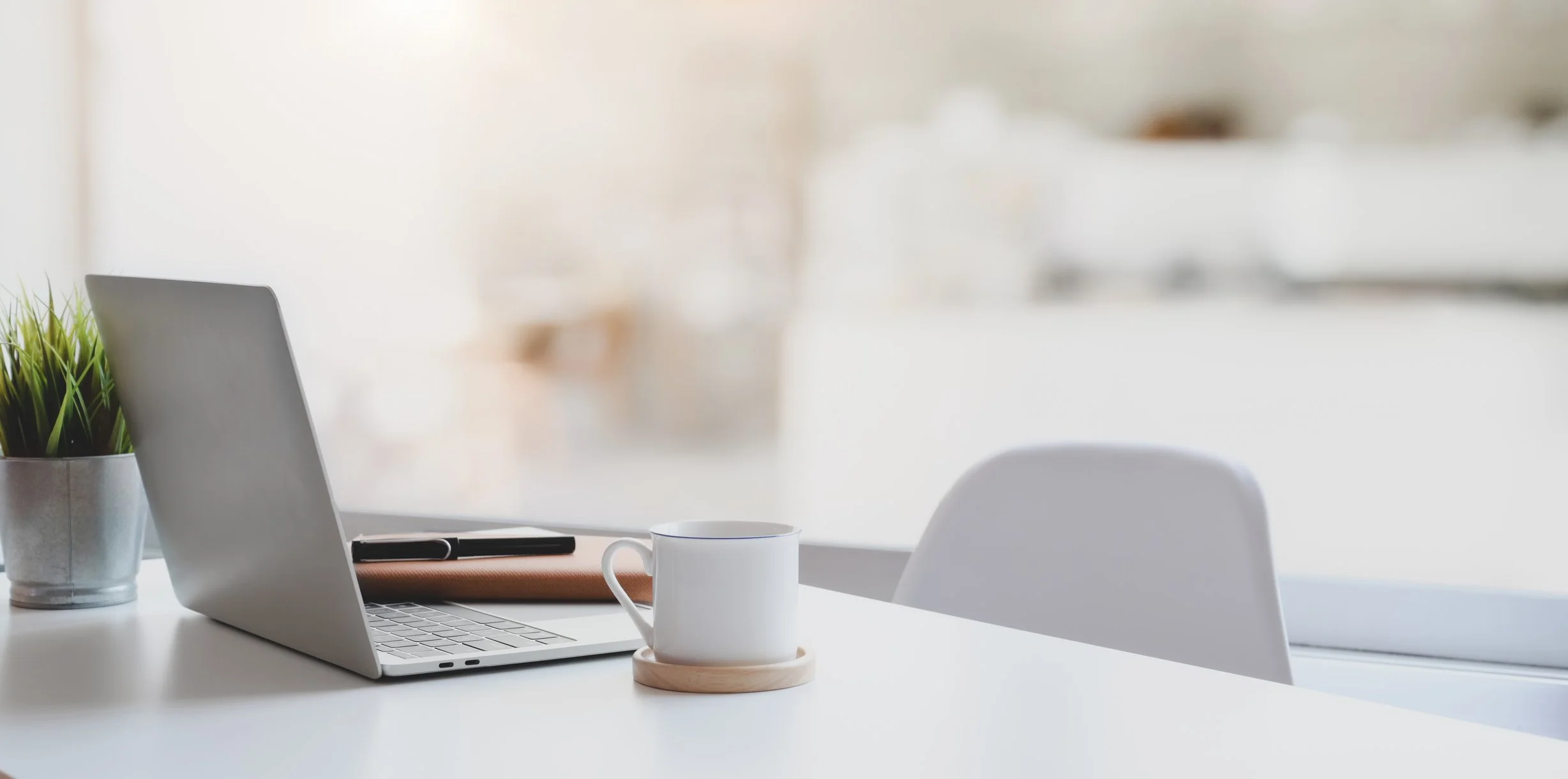 Modern office or workspace with a silver laptop, a white mug on a wooden coaster, a potted plant, a closed notebook, and a pen on a white table, with a blurred background.