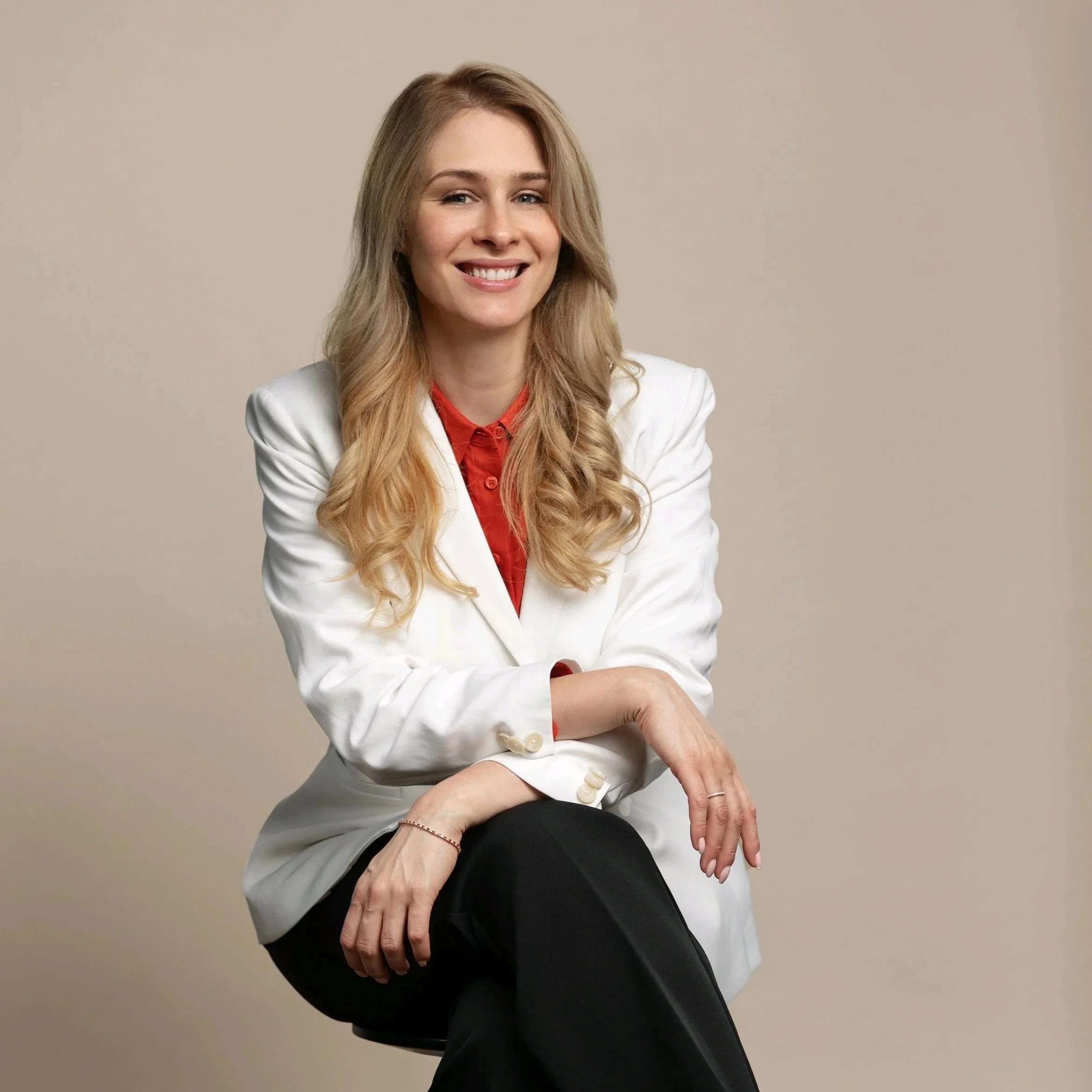 A young woman with long, wavy blonde hair, wearing a white blazer over a red shirt, sitting on a stool against a beige background, smiling.
