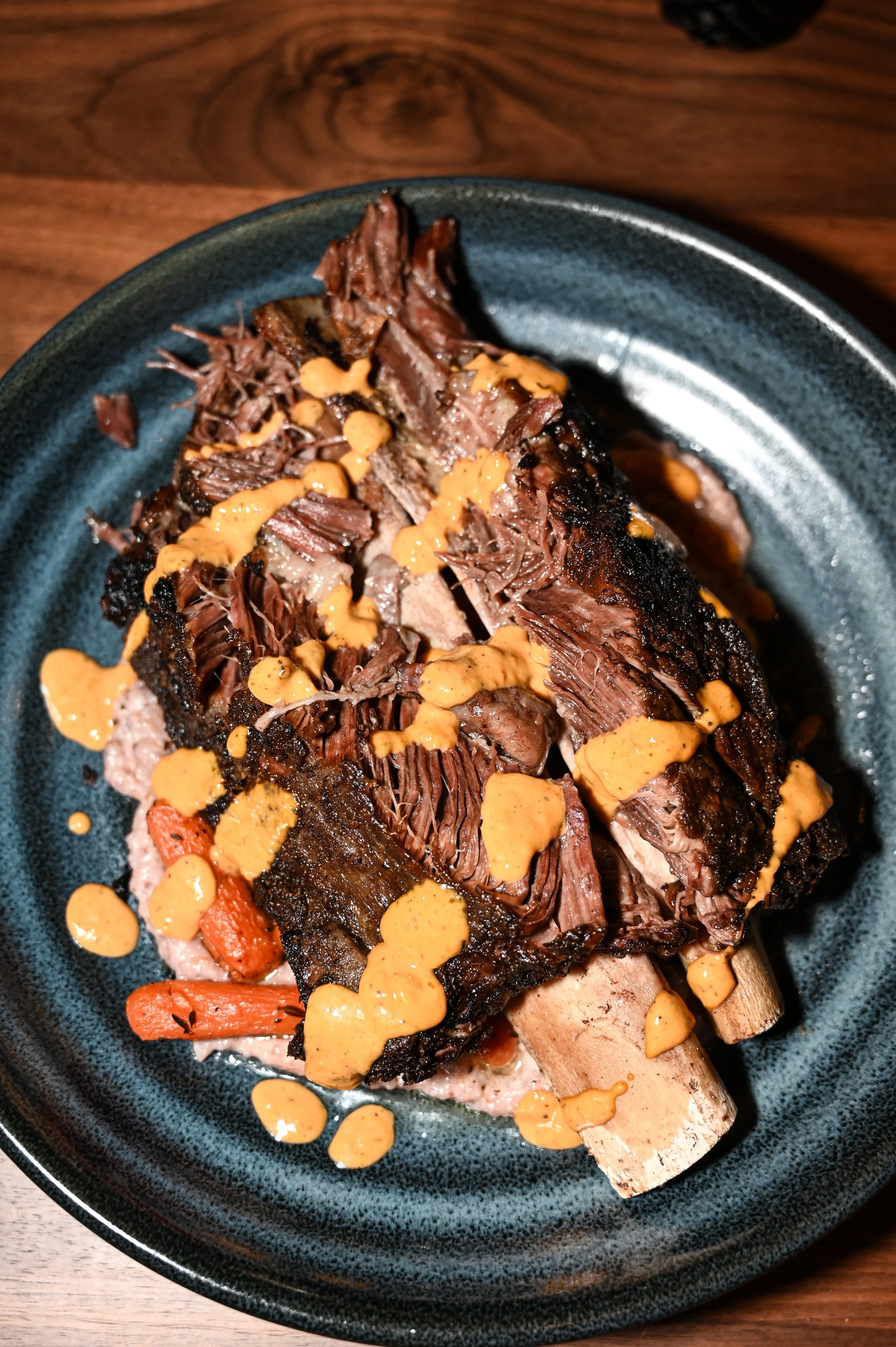A plate of shredded meat with orange sauce drizzled on top, with roasted carrots underneath, served on a dark ceramic plate on a wooden table.