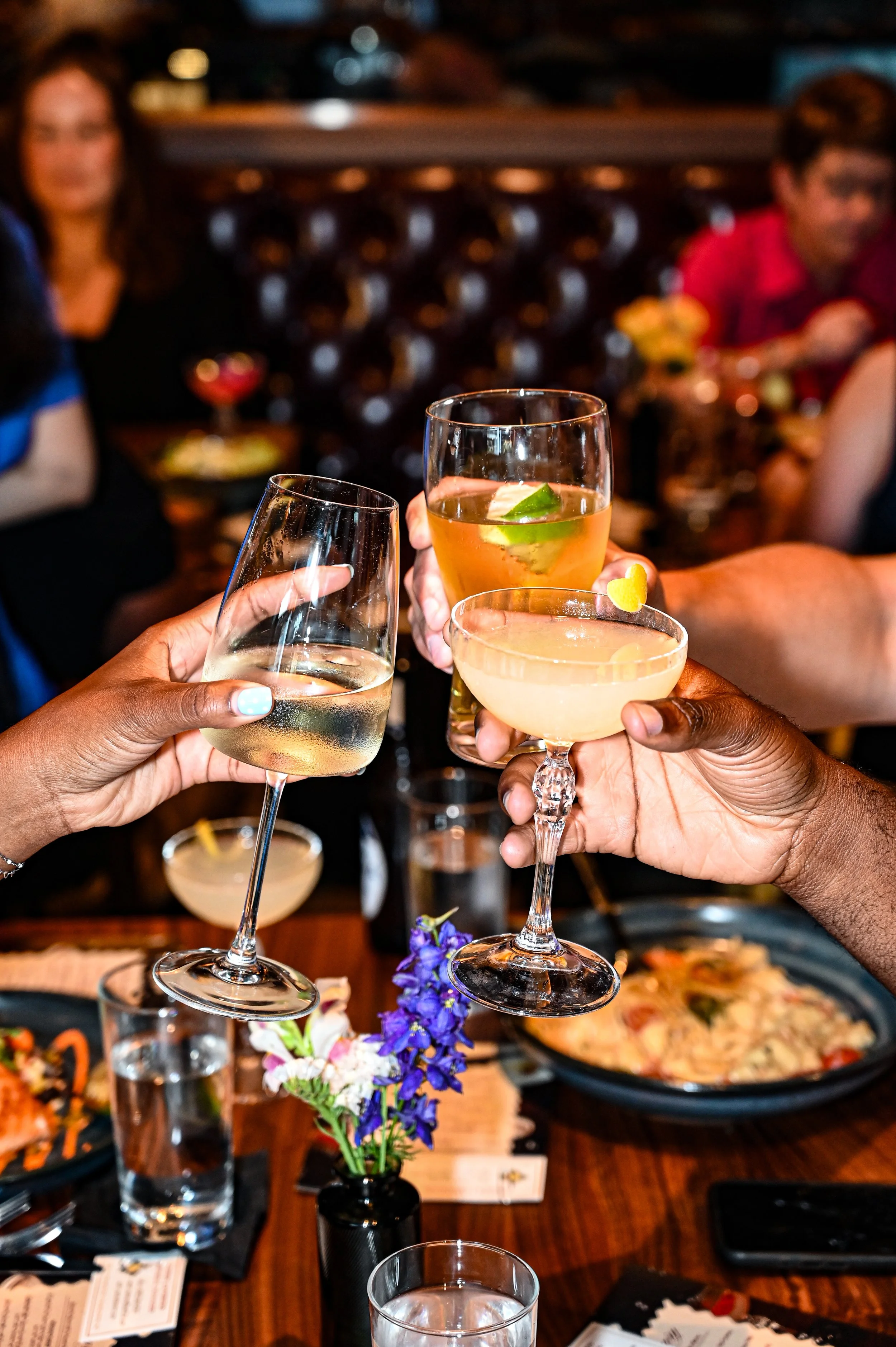 People raising glasses for a toast at a restaurant table with food and a vase of flowers in the center.