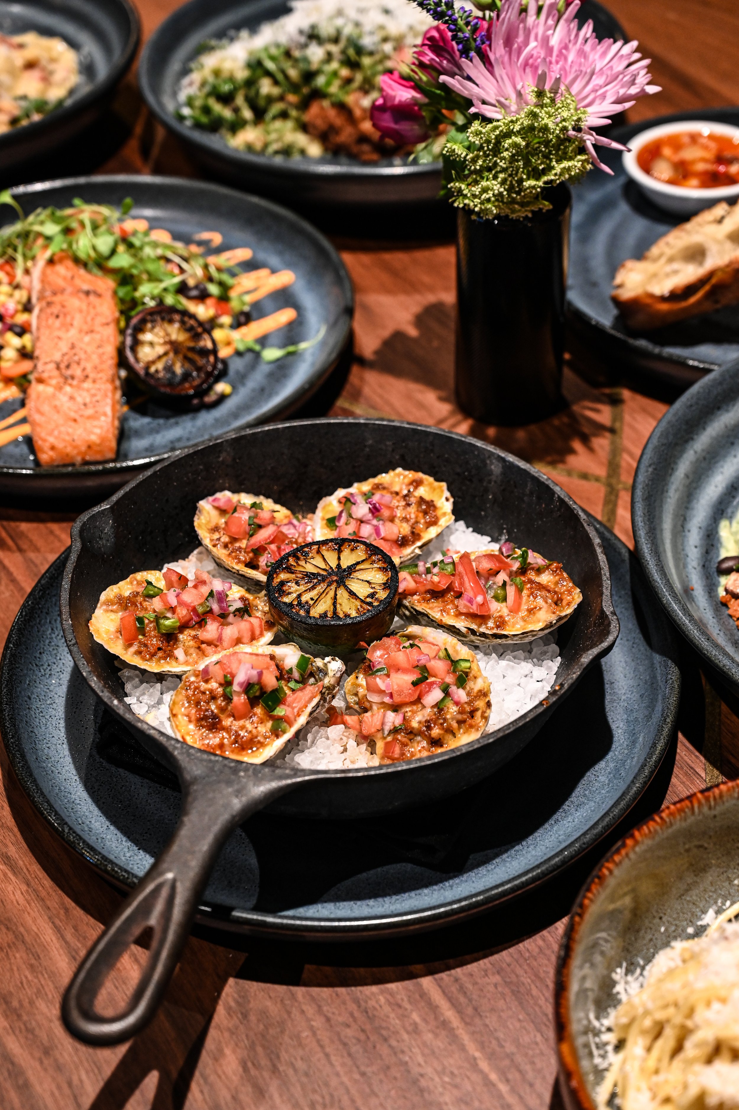 A cast iron skillet filled with baked oysters topped with diced tomatoes, green onions, and a grilled lemon slice in the center, on a bed of ice. Various plates of food and a small black vase with purple and pink flowers are on a wooden table in the background.