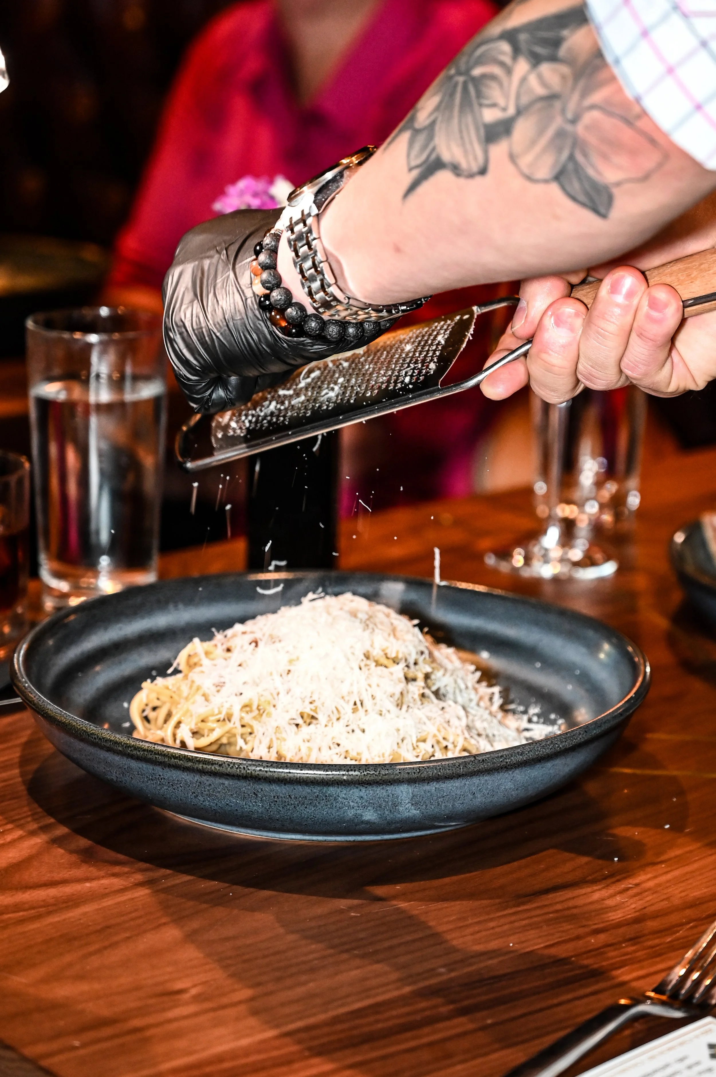 A plate of spaghetti topped with grated cheese on a wooden table, with a person's arm and hand using a cheese grater to add more cheese. The person's arm has tattoos of flowers, and they are wearing a watch and bracelets. There are drinks and glasses visible in the background.