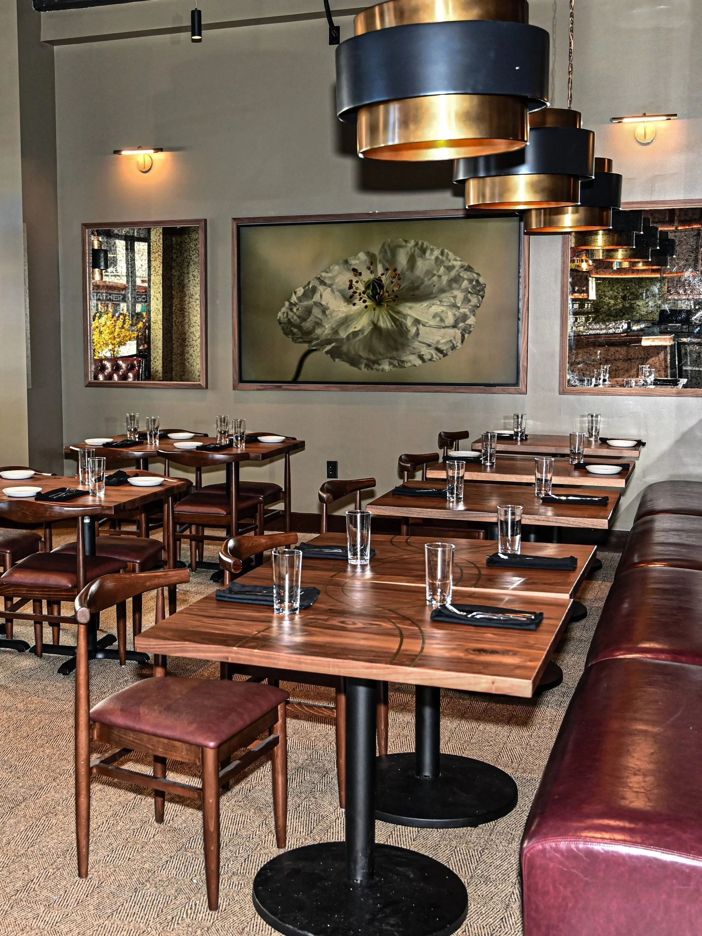 Interior view of a restaurant with wooden tables set with glasses and black napkins, leather chairs, decorative artwork on the walls, and modern hanging light fixtures.