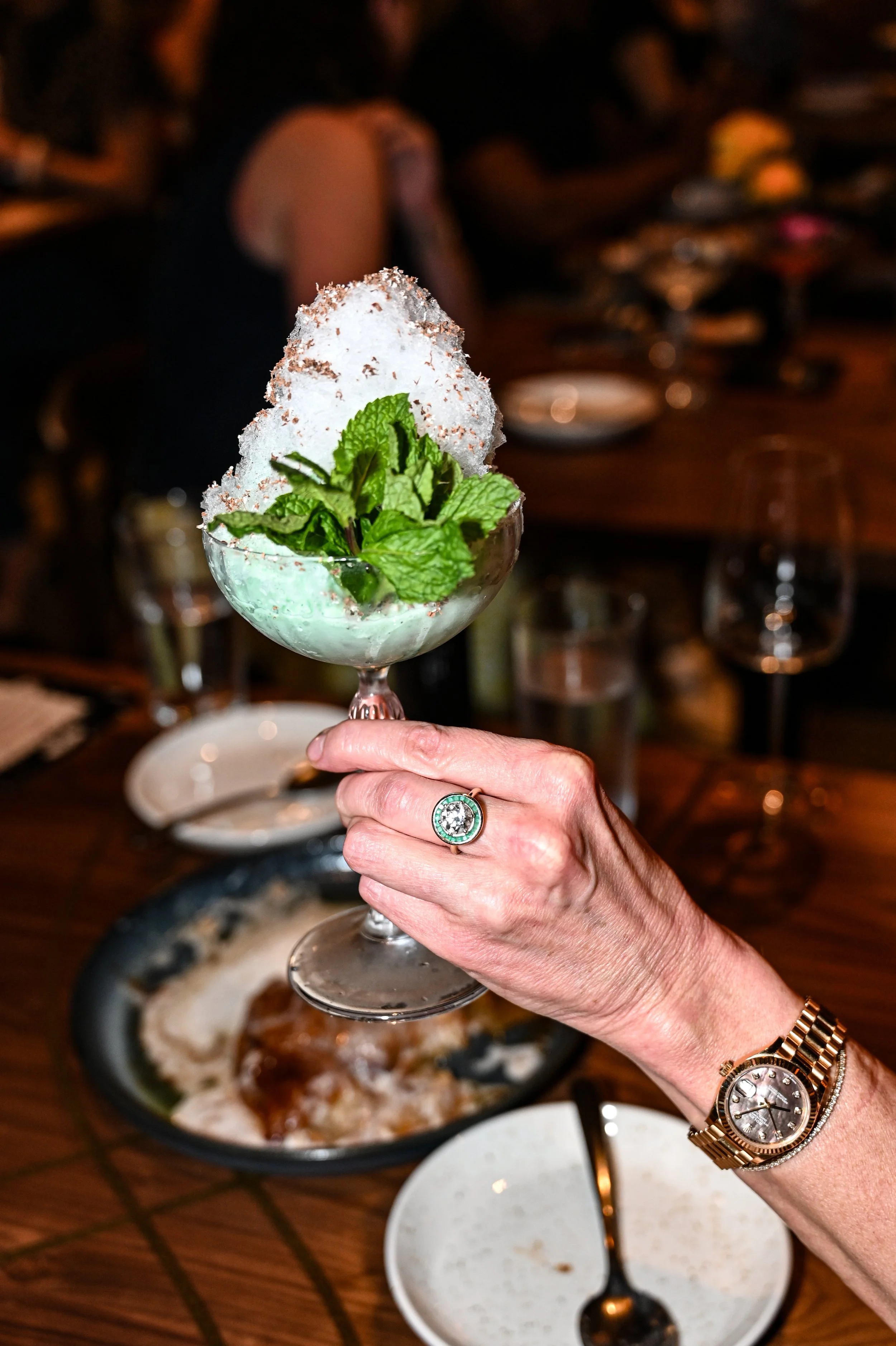 A woman's hand holding a cocktail glass with mint leaves and snow cone topped with chocolate shavings in a restaurant setting.